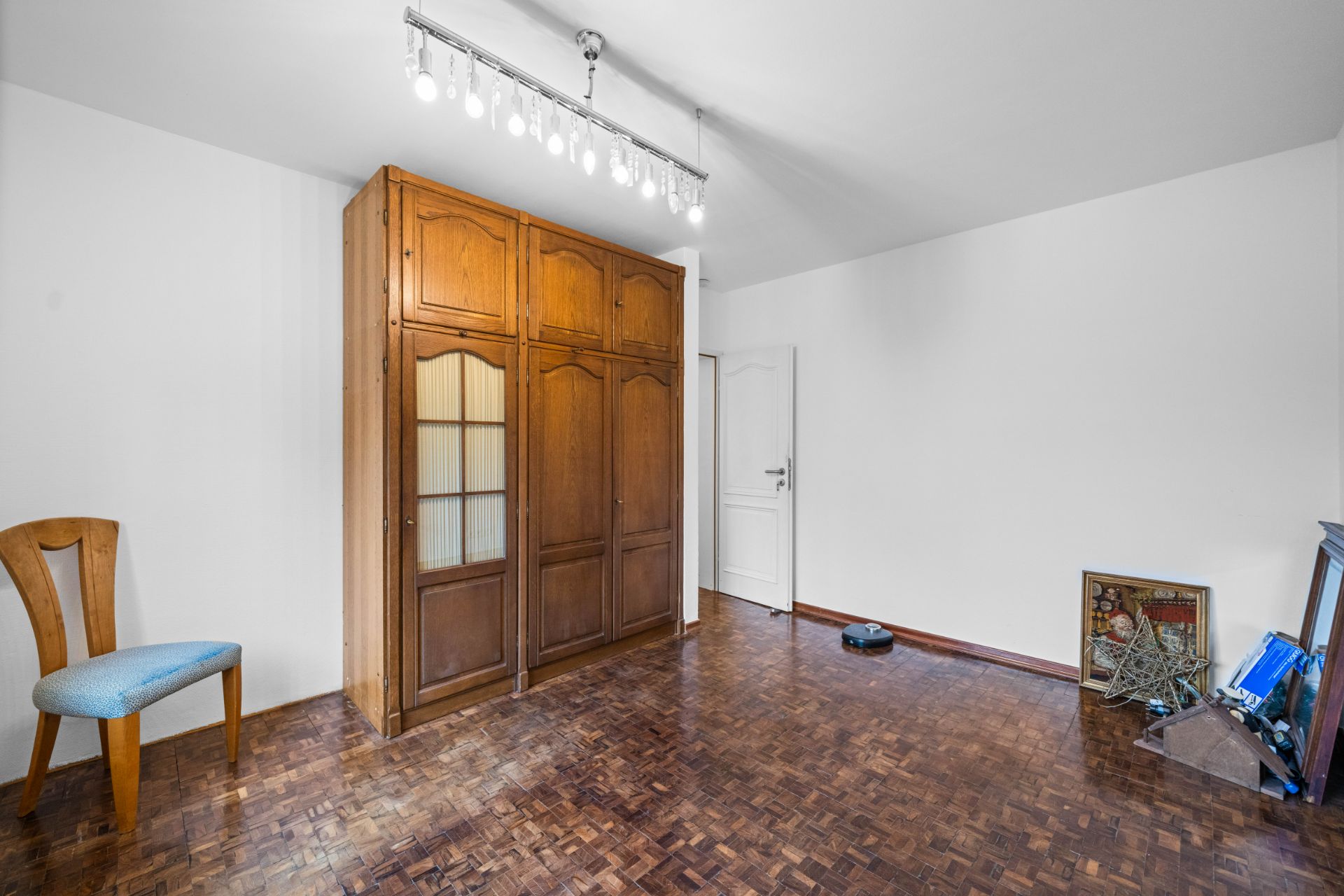 Leeres Zimmer mit Holzschrank, Stuhl und einigen Gegenständen am Boden.