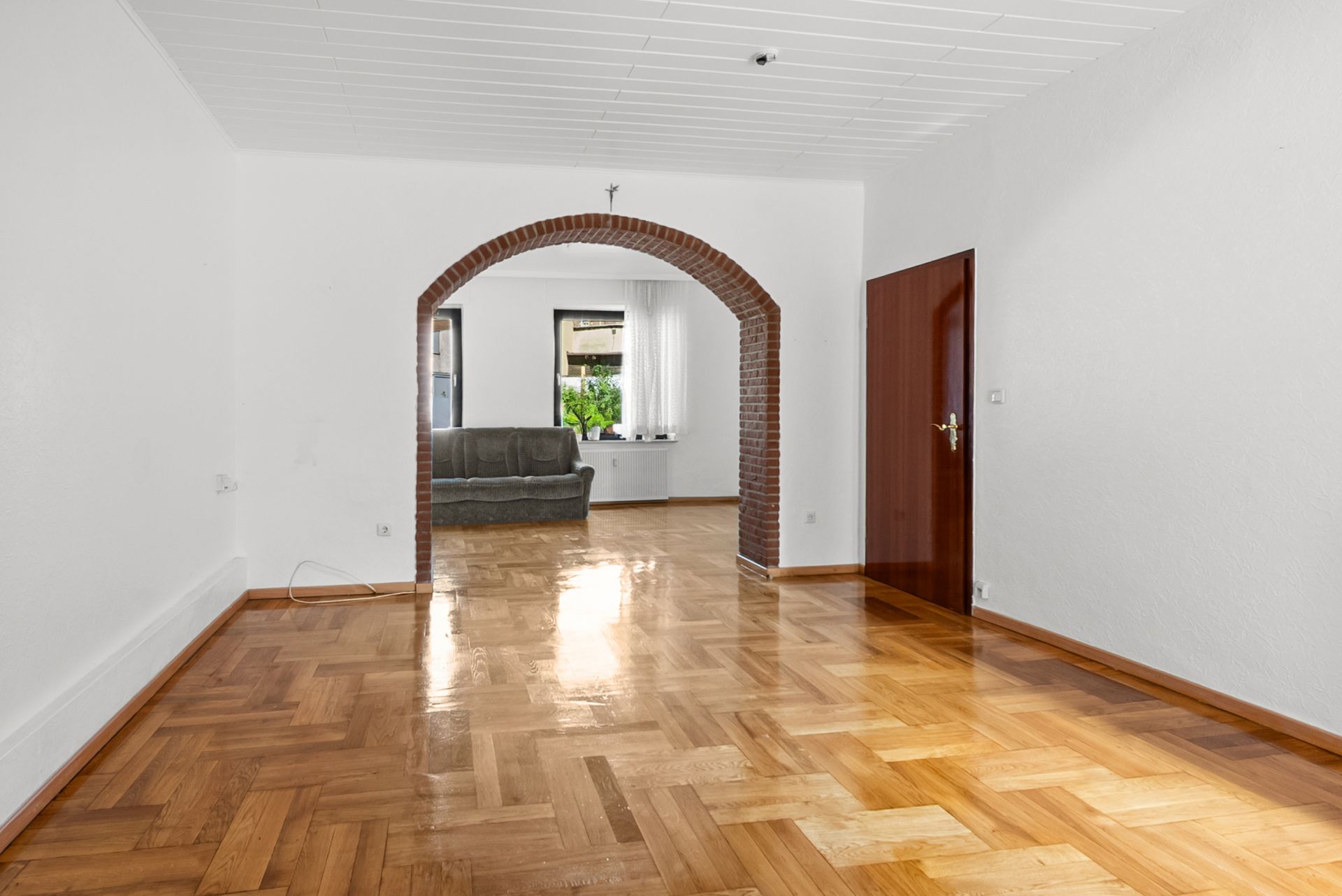 Leeres Wohnzimmer mit Parkettboden, einem gemauerten Rundbogen und Sofa im Hintergrund.