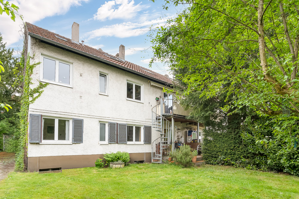 Zweistöckiges Wohnhaus mit Balkon, Außentreppe und Garten, umgeben von Bäumen.
