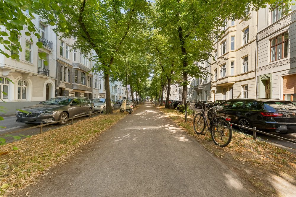 Baumbestandener Gehweg zwischen Wohnhäusern, auf beiden Seiten Autos und Fahrräder geparkt.