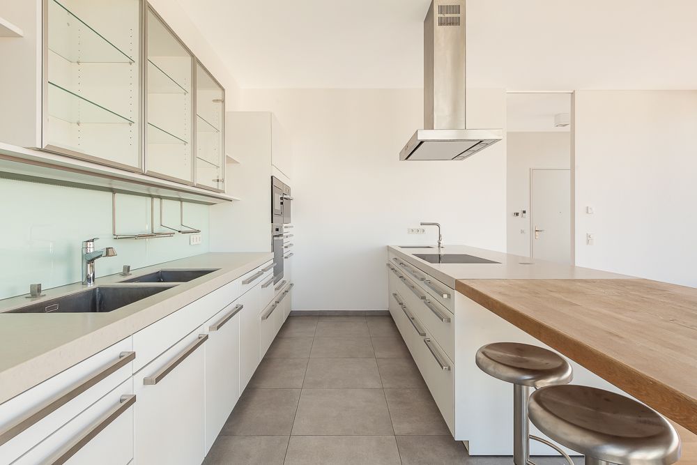 Moderne, helle Einbauküche mit Kochinsel, Edelstahlspüle und zwei Hockern am Tresen.