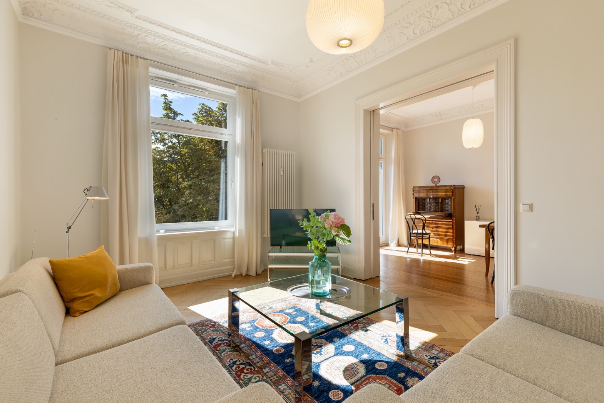 Helles Wohnzimmer mit Sofa, Glastisch, Teppich, großem Fenster und Blick in ein weiteres Zimmer.