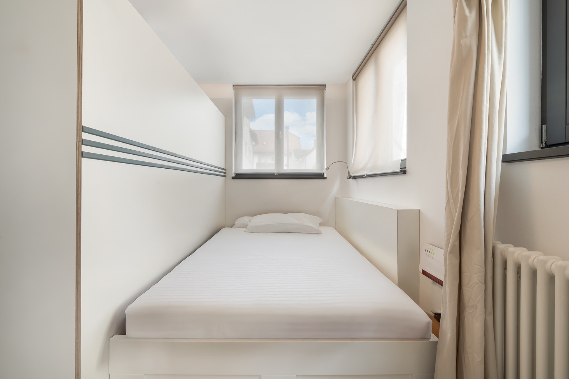 Kleines Schlafzimmer mit Einzelbett, weißer Bettwäsche und Fenstern auf zwei Seiten.