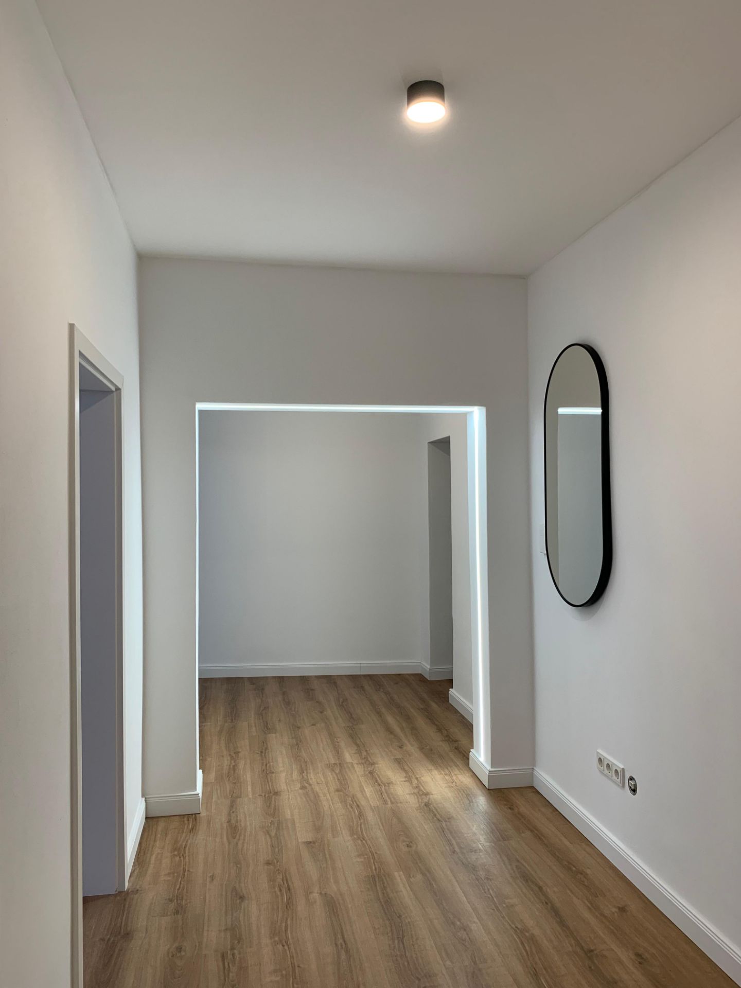 Heller Flur mit Holzfußboden, weißer Wand und ovalem Spiegel an der rechten Wand.