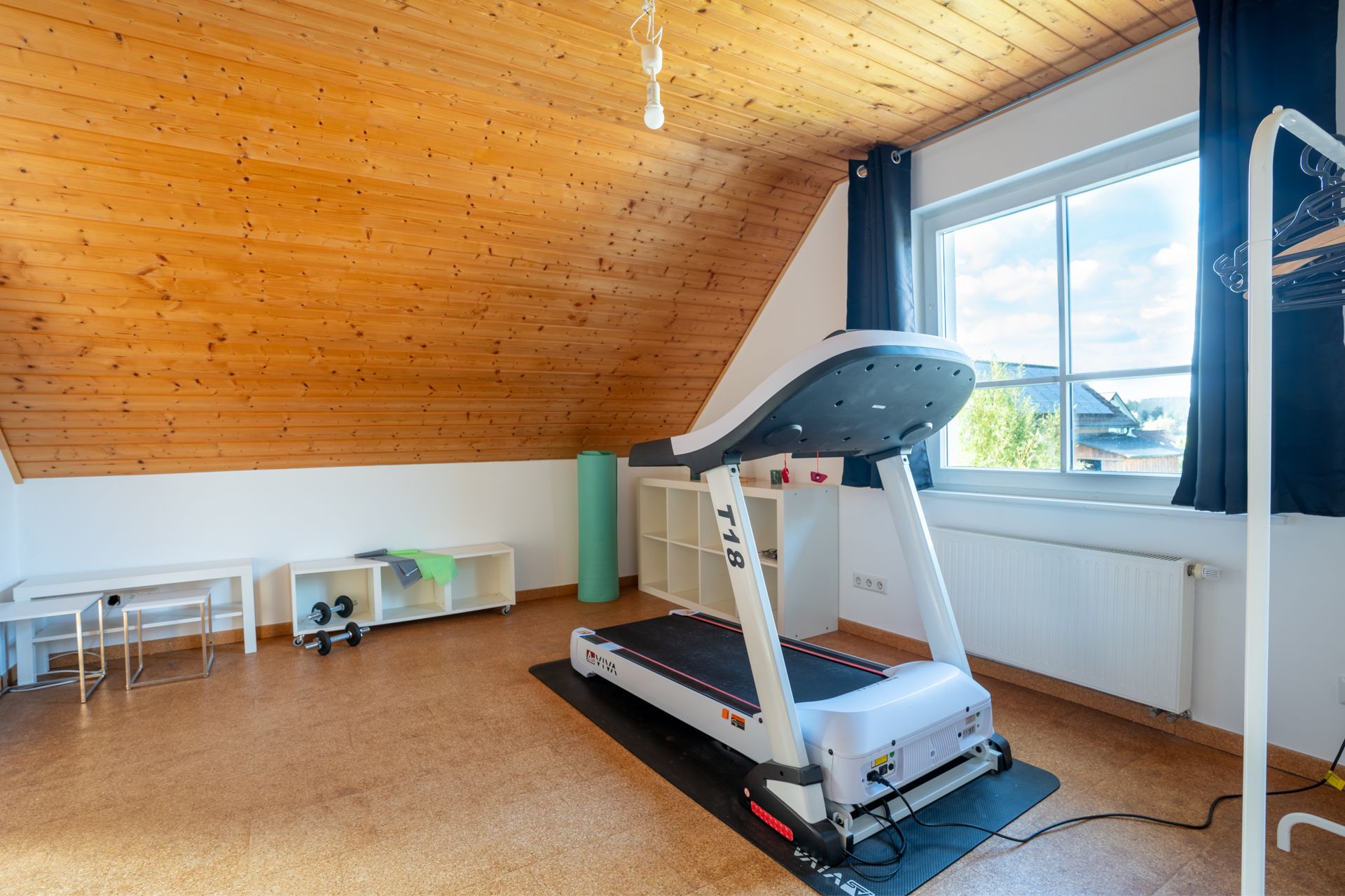 Heller Raum mit Dachschräge, Laufband, Regal, Fenster und Sportzubehör.