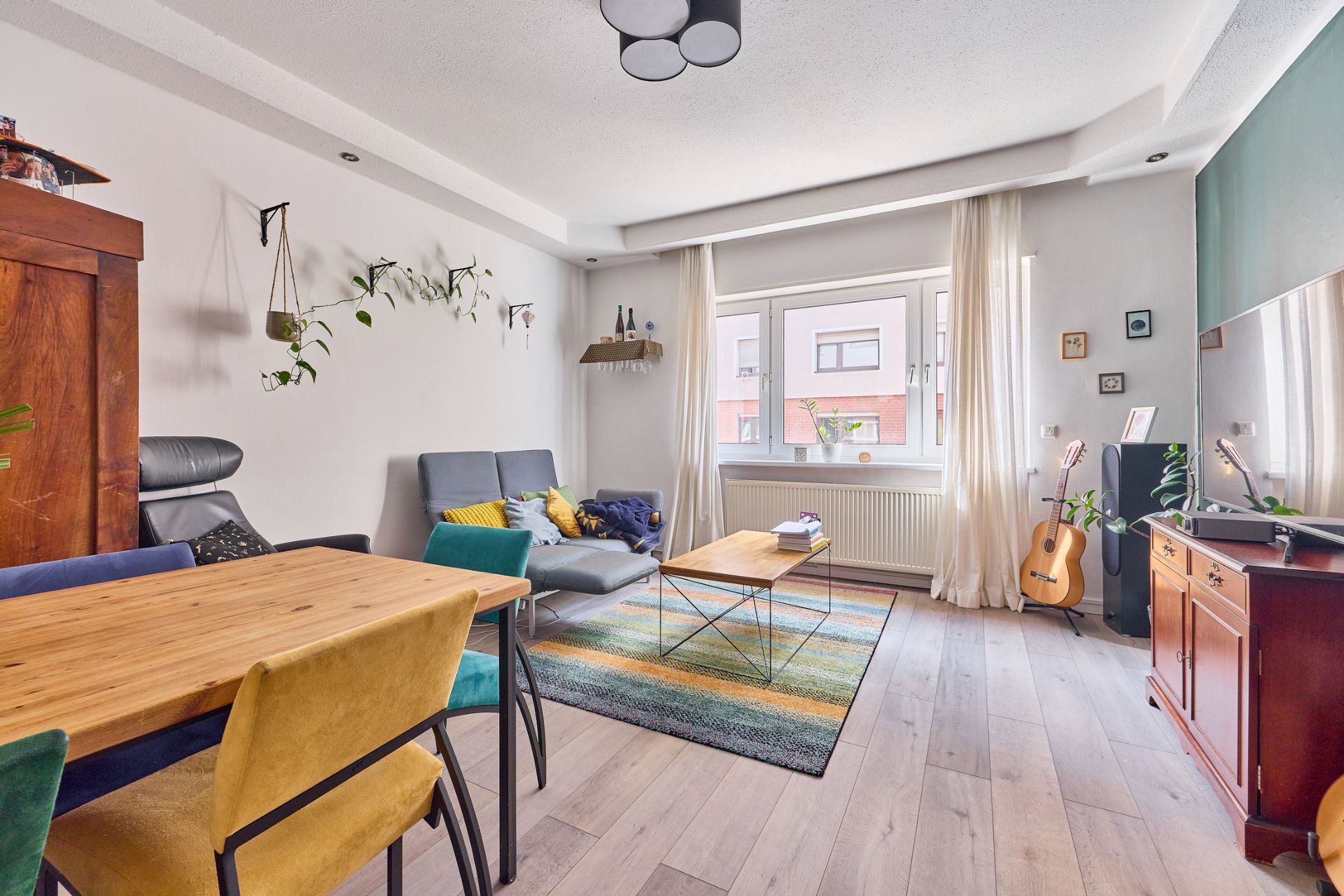 Heller Wohnraum mit Sofa, Esstisch, bunten Stühlen, Regal, Gitarre und großem Fenster.