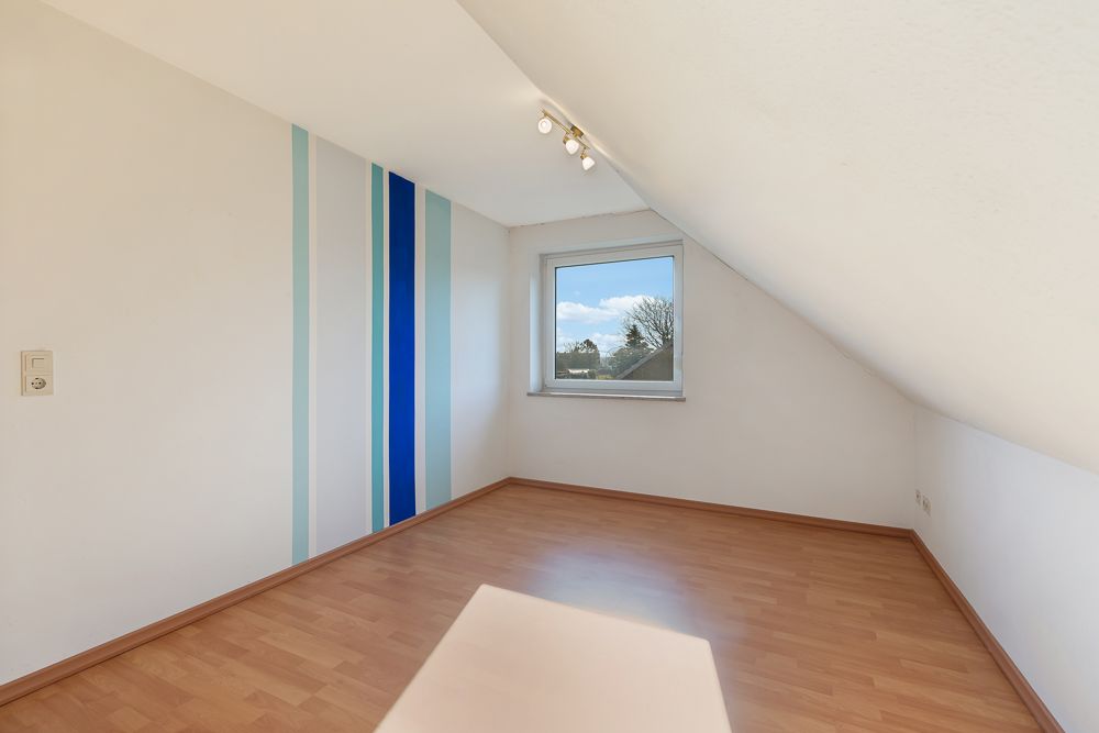 Leeres Zimmer mit Dachschräge, Holzboden und einem Fenster, eine Wand mit blauen Streifen.