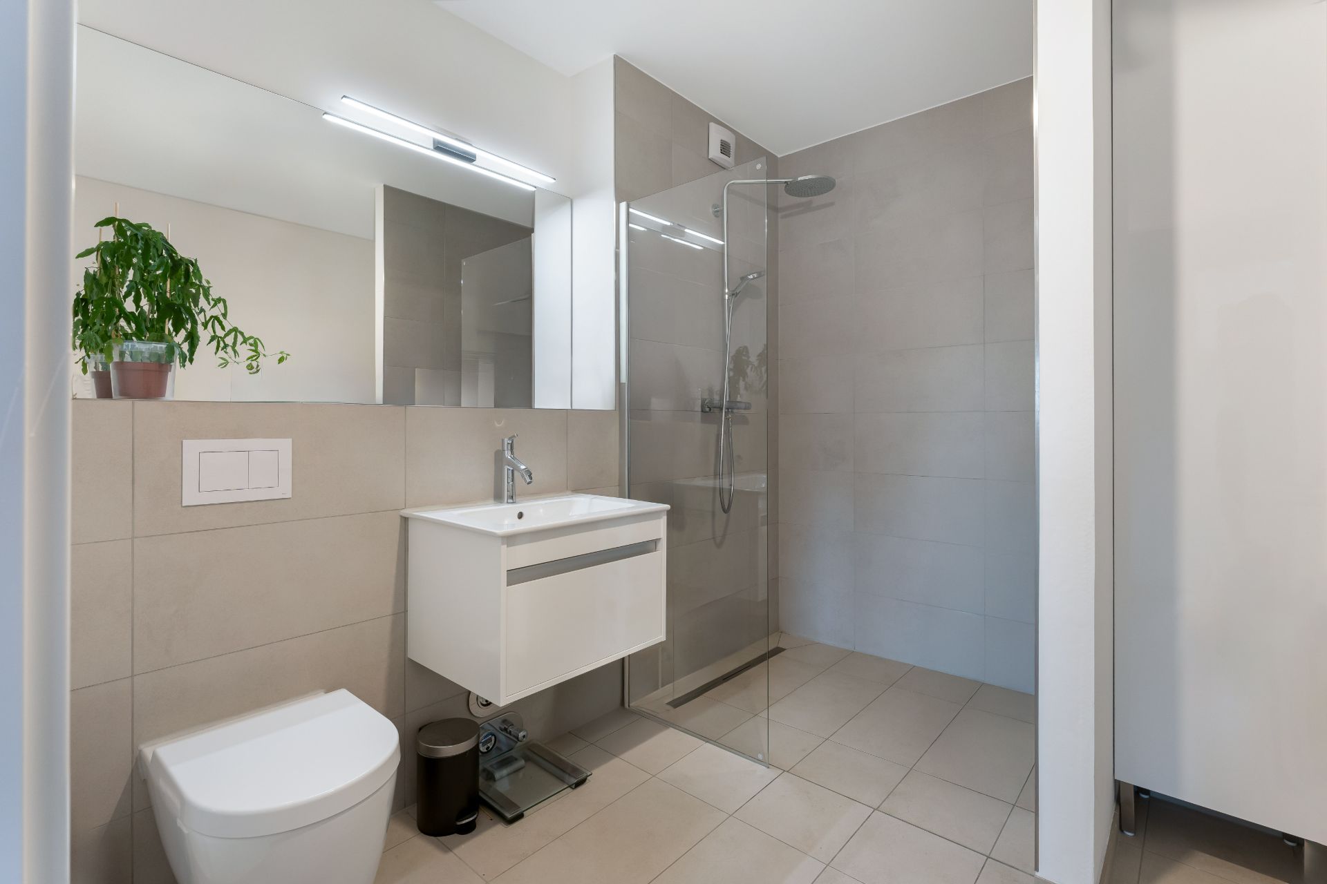 Modernes Badezimmer mit Toilette, Waschbecken, Glasdusche und einer Pflanze auf dem Regal.