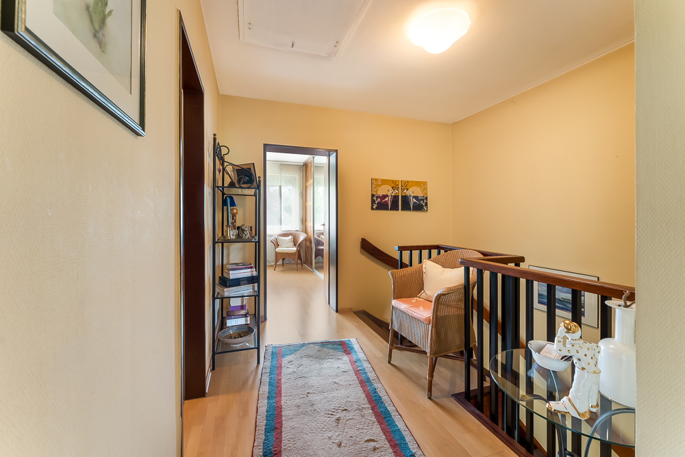 Heller Flur mit Teppich, Treppengeländer, Regal, Stuhl und Blick in ein weiteres Zimmer.
