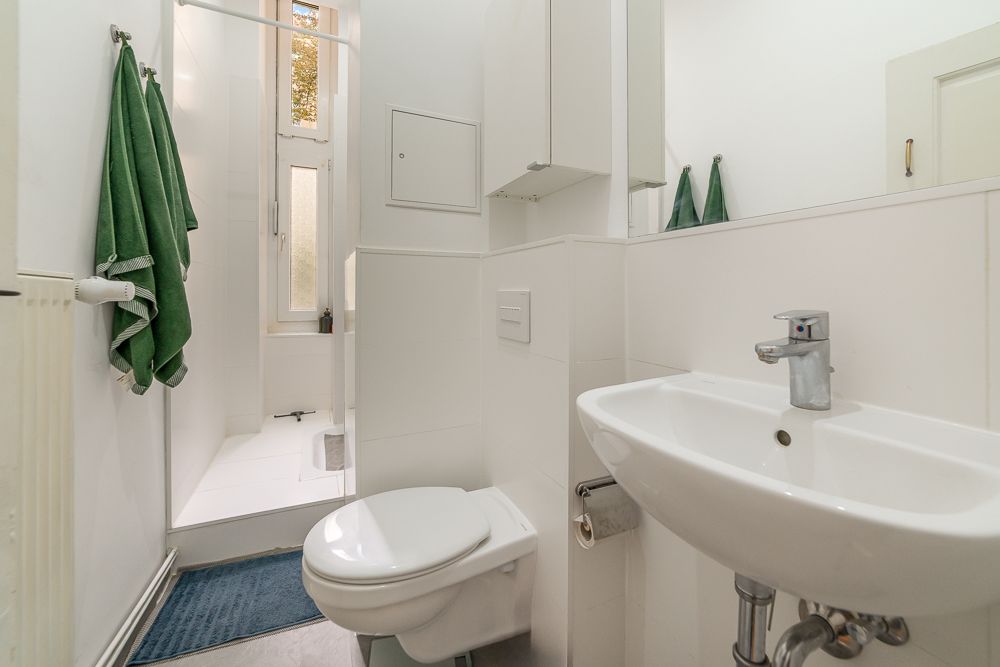 Kleines Badezimmer mit Toilette, Waschbecken, Dusche, Spiegel und zwei grünen Handtüchern.