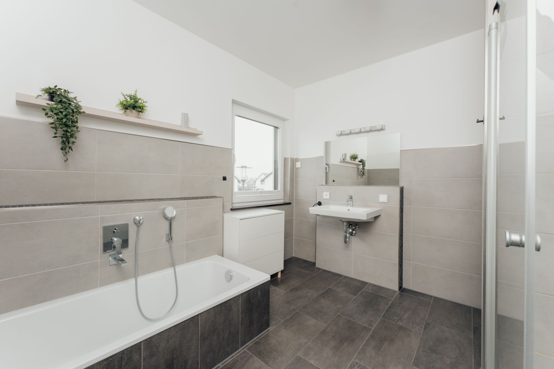 Modernes Badezimmer mit Badewanne, Waschbecken, Spiegel und grauen Fliesen.