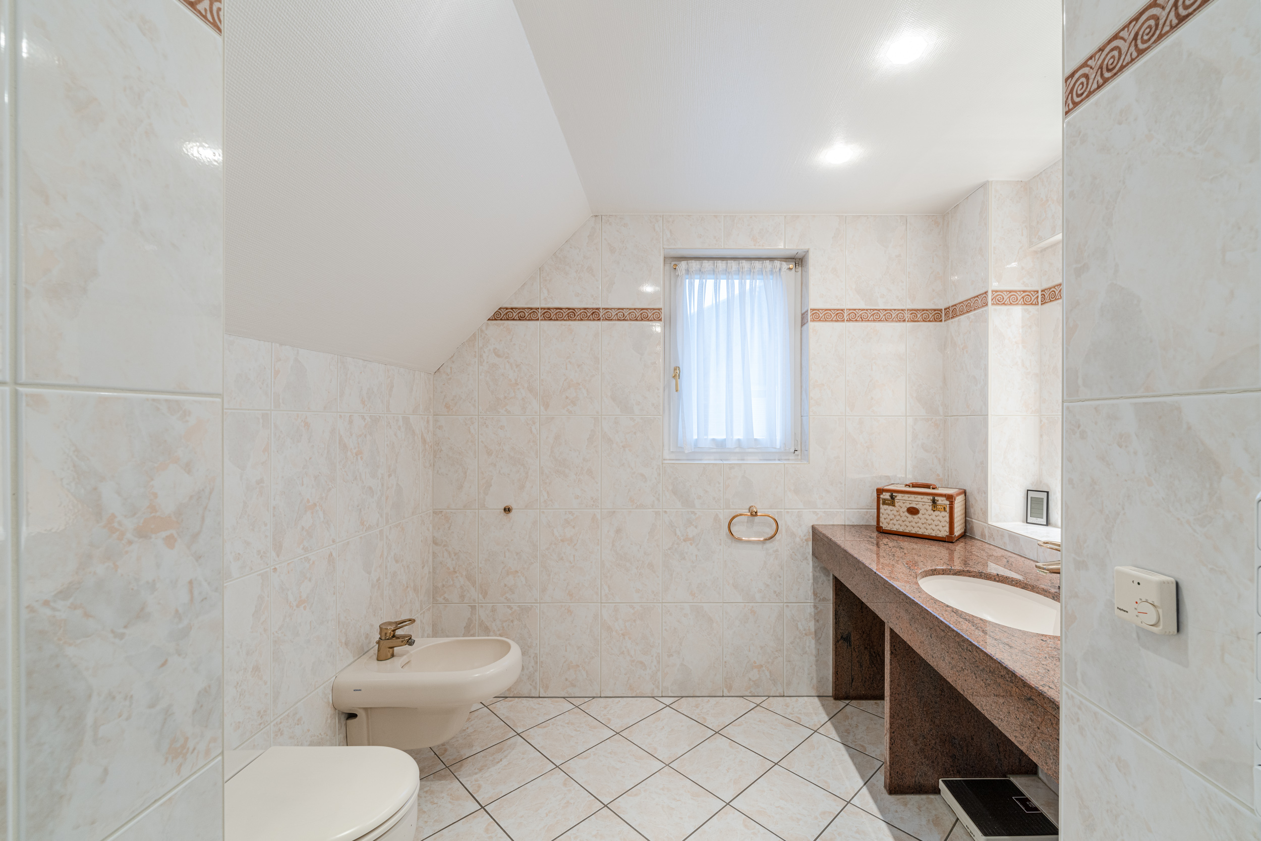 Helles Badezimmer mit Bidet, WC, Waschbecken und Fenster, gefliest mit Marmoroptik.