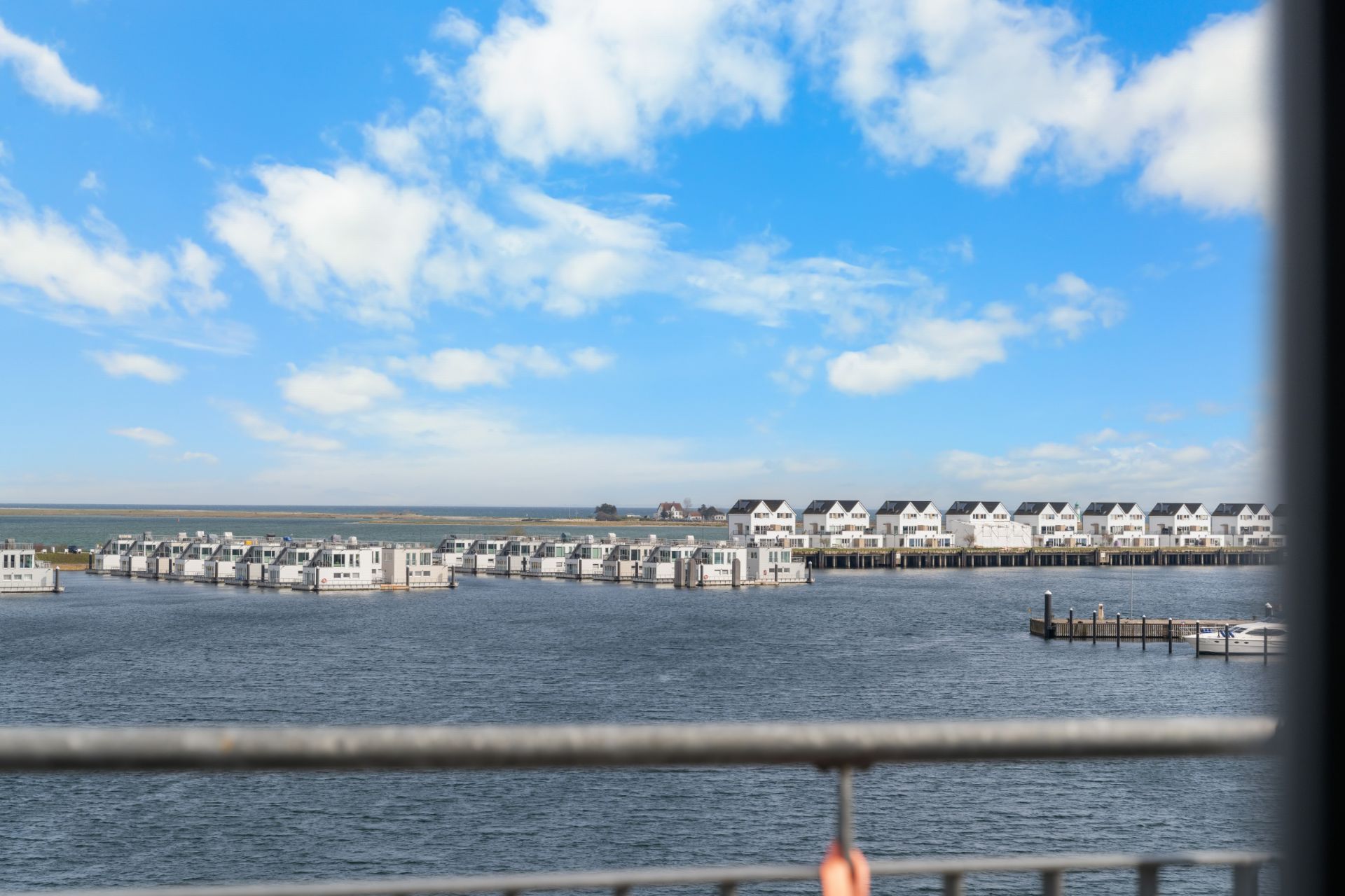 Blick über ein Geländer auf Wasser und weiße Häuser am Ufer unter blauem Himmel.