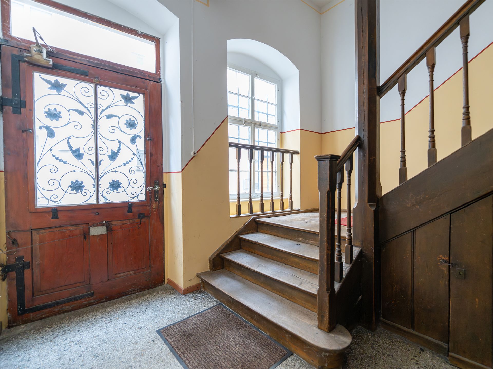 Eingangsbereich mit Holztreppe, roter Tür mit Gittermuster und großem Fenster.