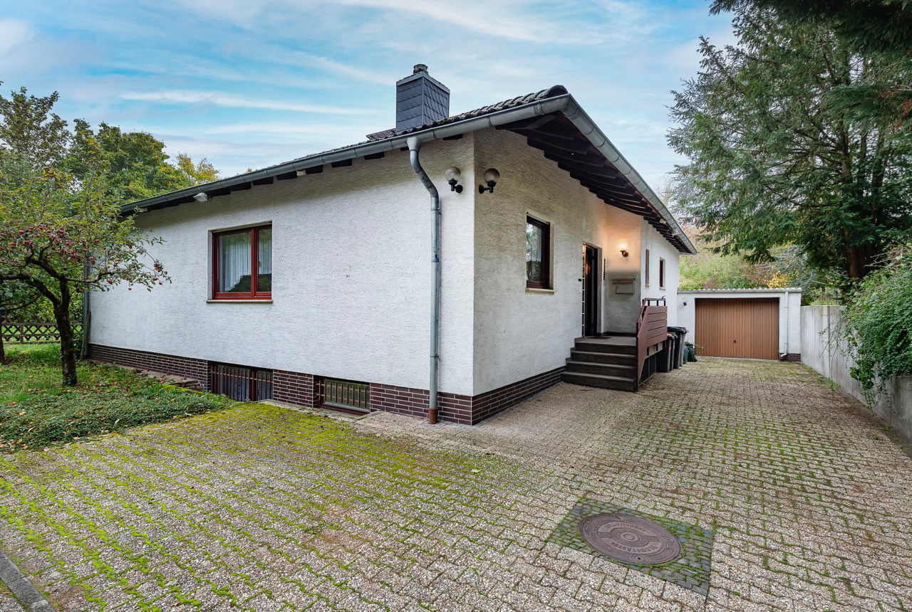 Weißes Einfamilienhaus mit Garage, gepflasterter Einfahrt und Garten mit Baum.