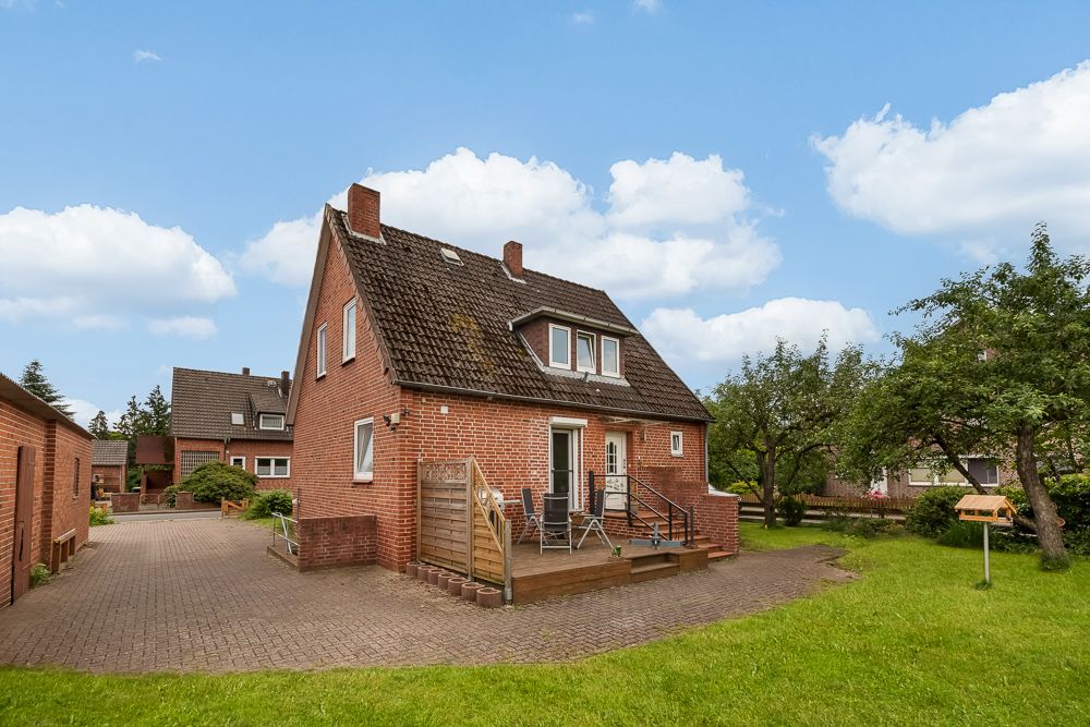 Backstein-Einfamilienhaus mit Terrasse und Garten, umgeben von Bäumen unter blauem Himmel.