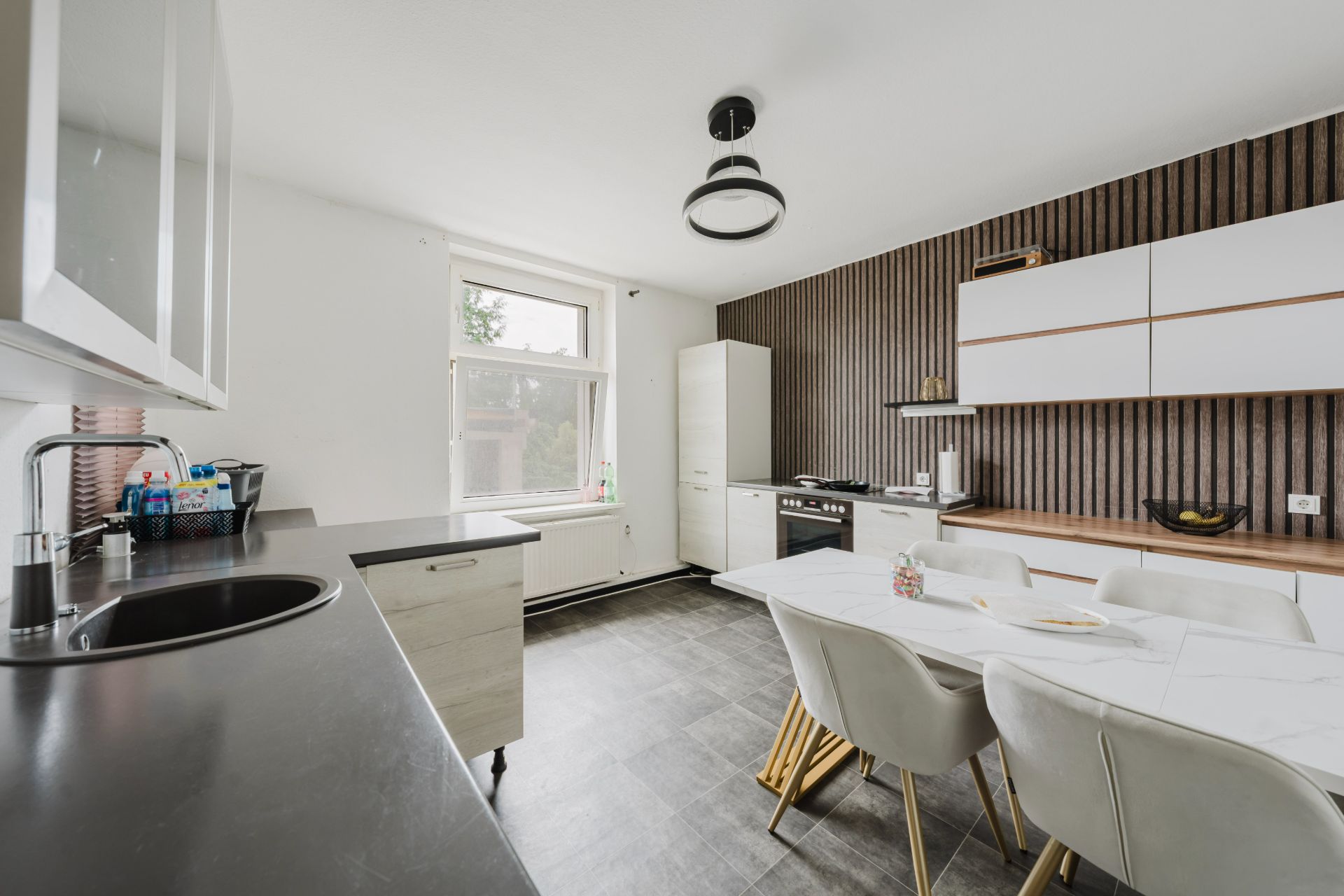Helle Küche mit Esstisch, modernen Stühlen und Geräten vor einer Wand mit Holzpaneelen.