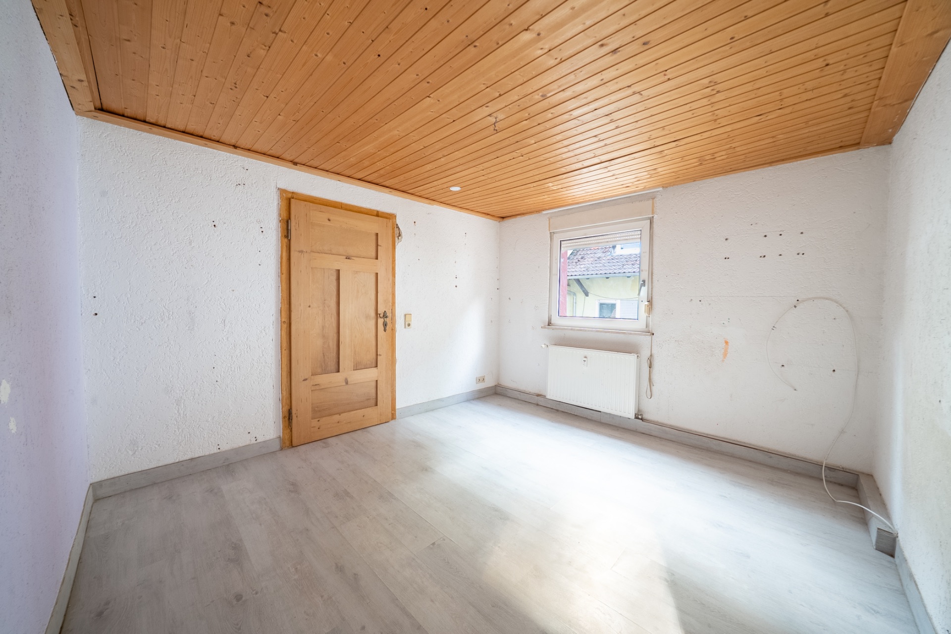 Leeres Zimmer mit Holztür, Fenster, weißem Heizkörper und Holzdecke.