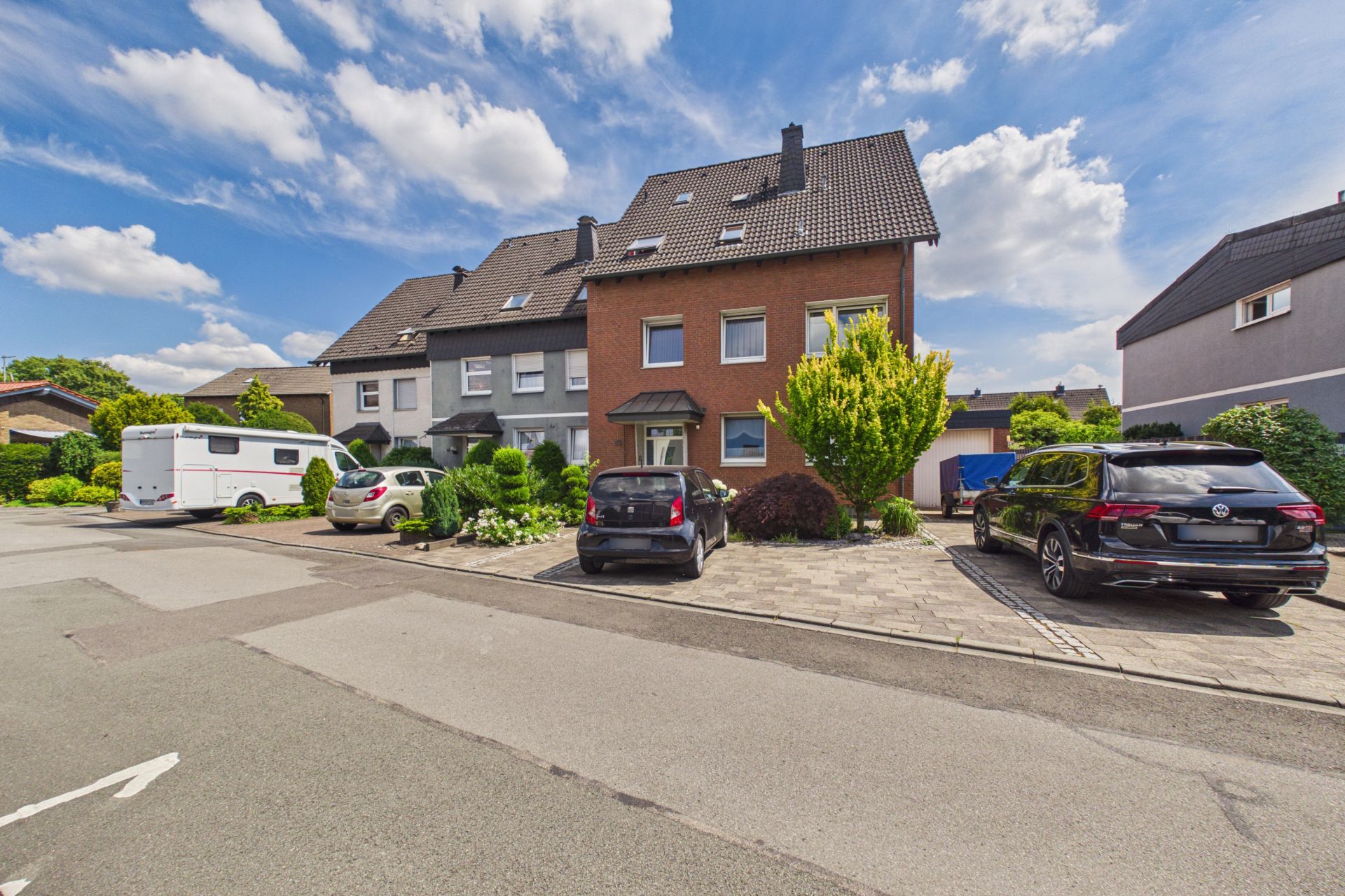 Reihenhaus mit Vorgarten und mehreren geparkten Autos auf einer Wohnstraße bei Sonnenschein.