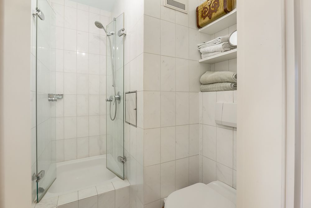 Modernes Bad mit Dusche, weißen Fliesen, WC und offenen Regalen mit Handtüchern.