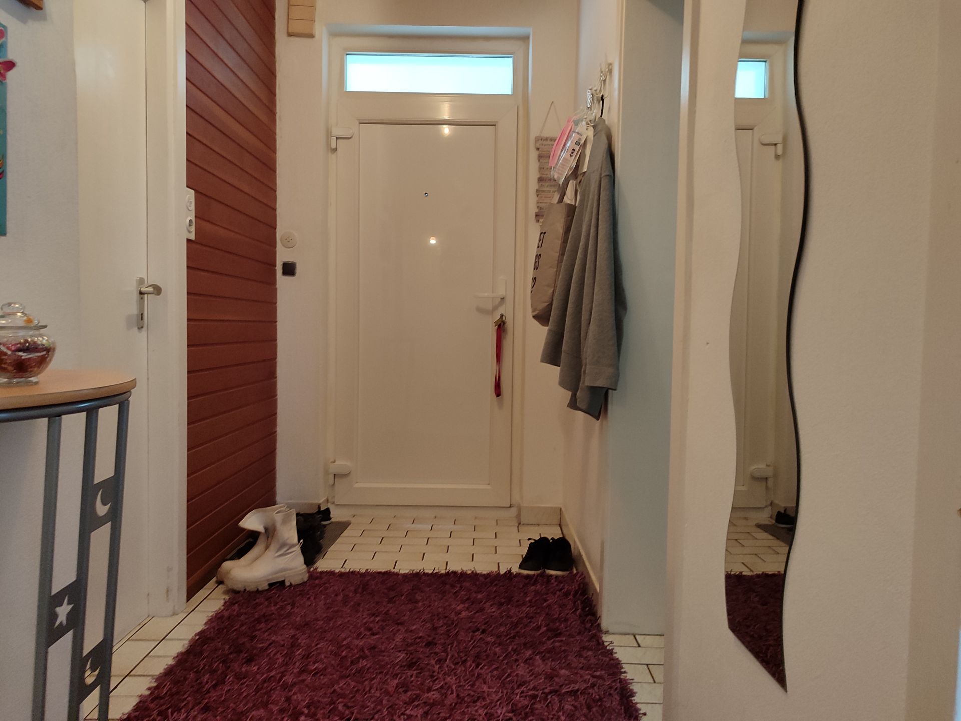 Flur mit weißer Haustür, lila Teppich, Schuhen und Jacken an Haken.