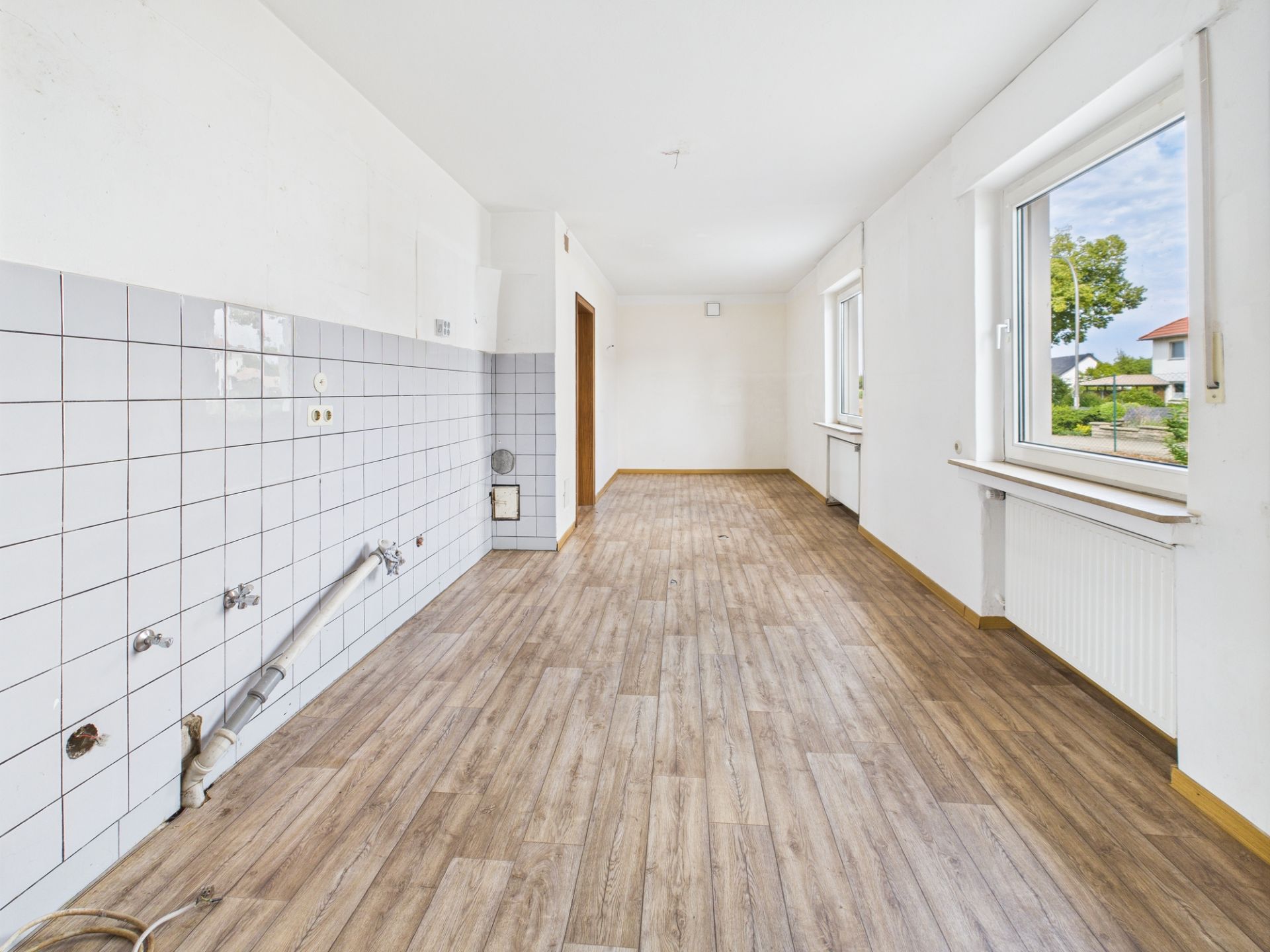 Leerer Raum mit weißen Fliesen an der Wand, zwei Fenstern und Holzboden.