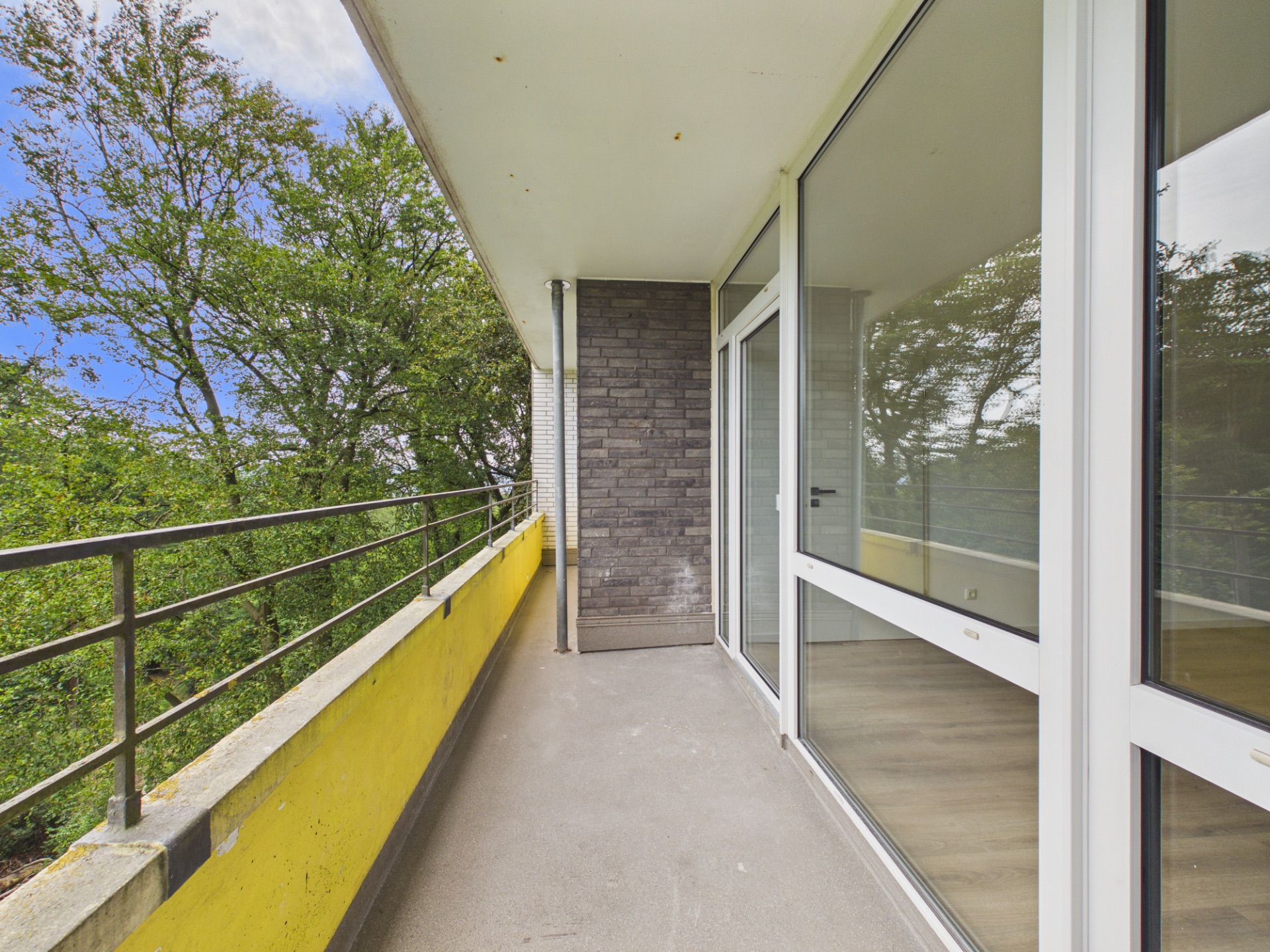 Balkon mit gelbem Geländer und Blick auf Bäume, angrenzend an Glastüren.