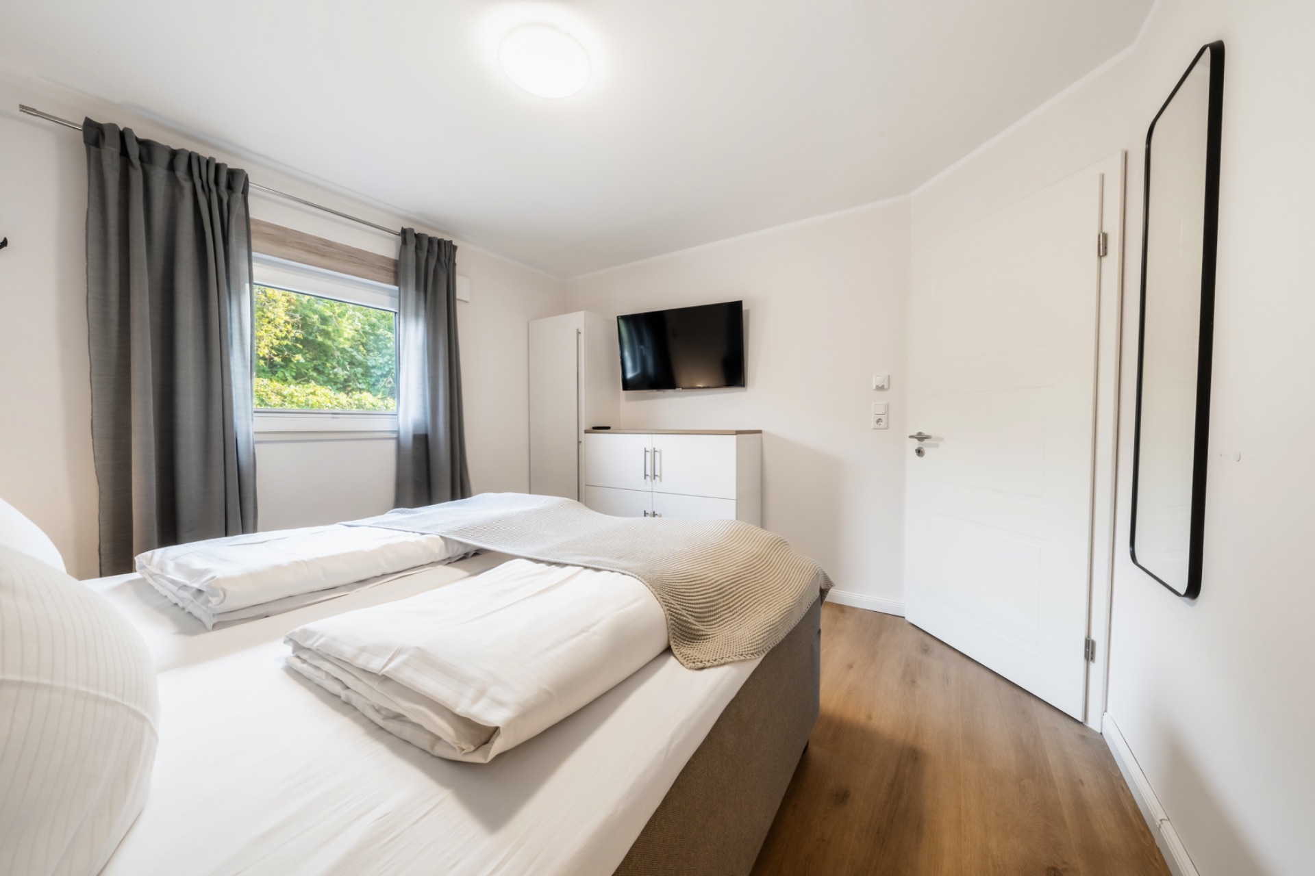 Helles Schlafzimmer mit Doppelbett, Fernseher, Schrank, Spiegel und Fenster mit grauen Vorhängen.