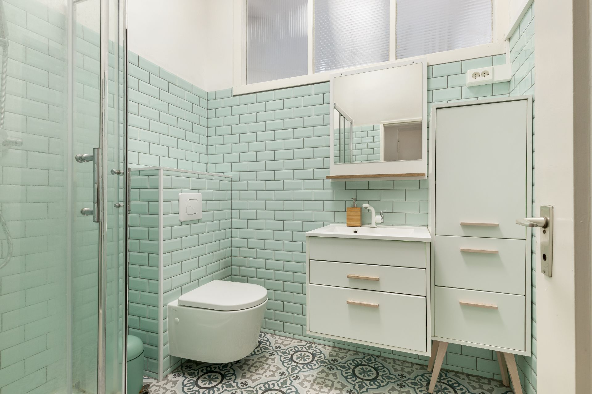 Helles Bad mit grünen Fliesen, Dusche, Hängetoilette, Waschbecken und Schrank.