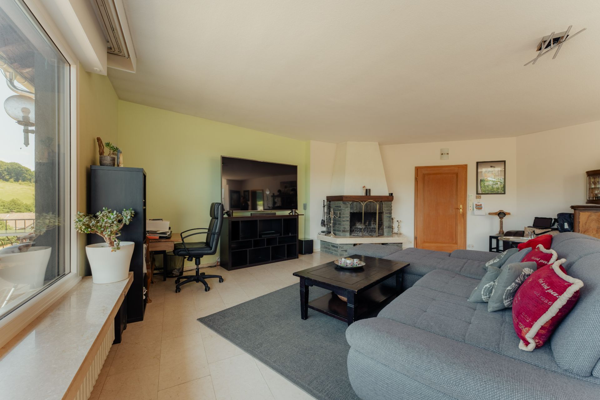 Wohnzimmer mit grauem Sofa, Kamin, großem Fenster, Schreibtisch und Fernseher.