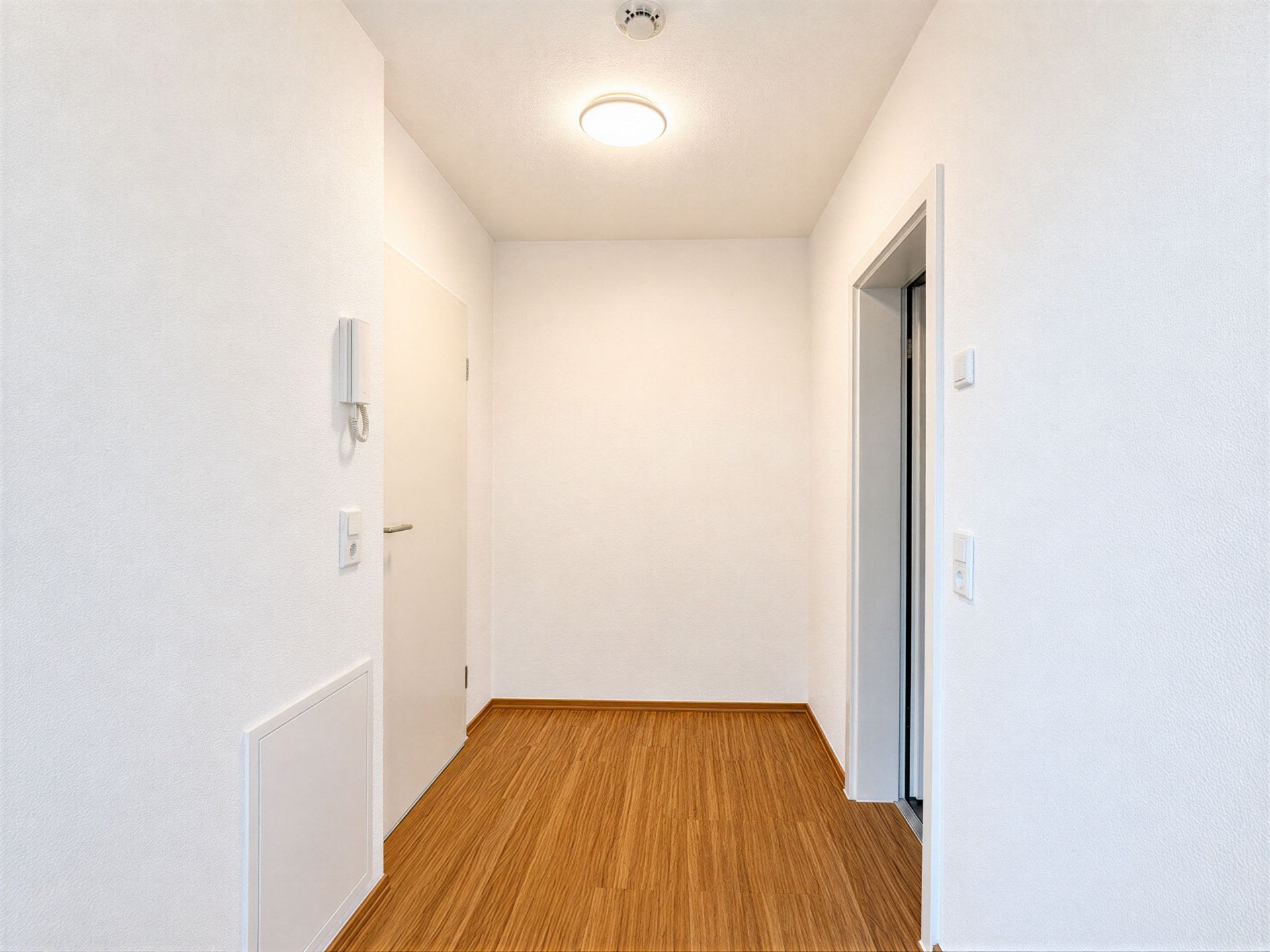 Heller Flur mit Holzboden, einer Tür links und einer offenen Tür rechts.