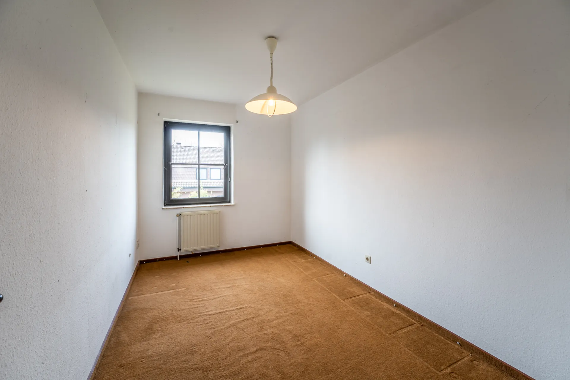 Leeres Zimmer mit braunem Teppich, weißer Wand, Fenster und Deckenlampe.