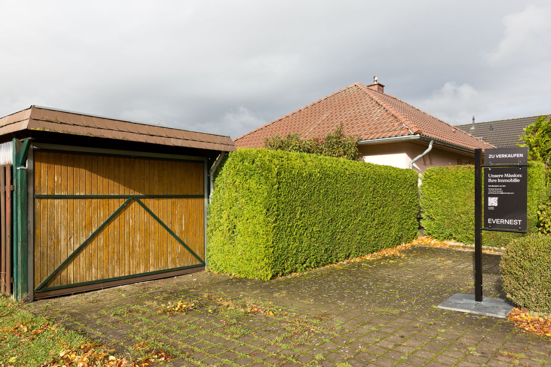 Garage mit Holztor, gepflasterte Einfahrt, grüne Hecke und Haus mit rotem Dach im Hintergrund.