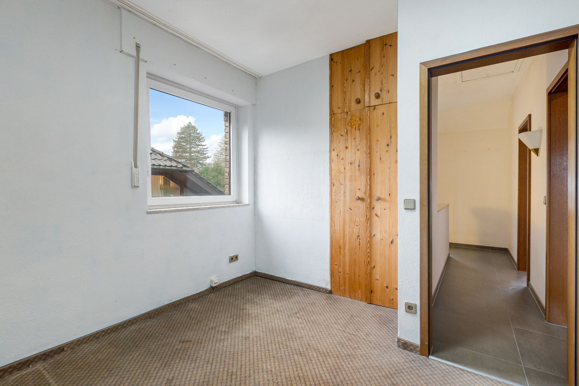 Kleines Zimmer mit Teppichboden, Fenster, Einbauschrank aus Holz und Blick auf einen Flur.