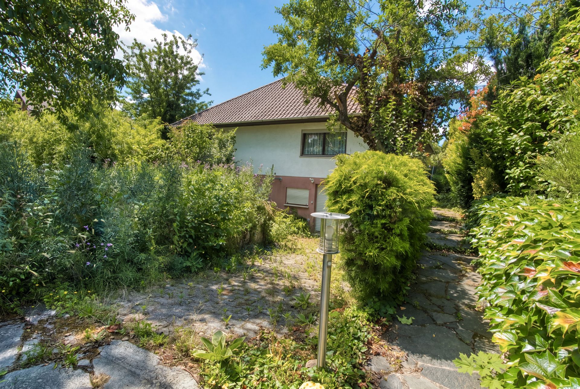 Haus mit Ziegeldach, von Bäumen und Sträuchern umgeben. Ein Steinweg führt durch den Garten.