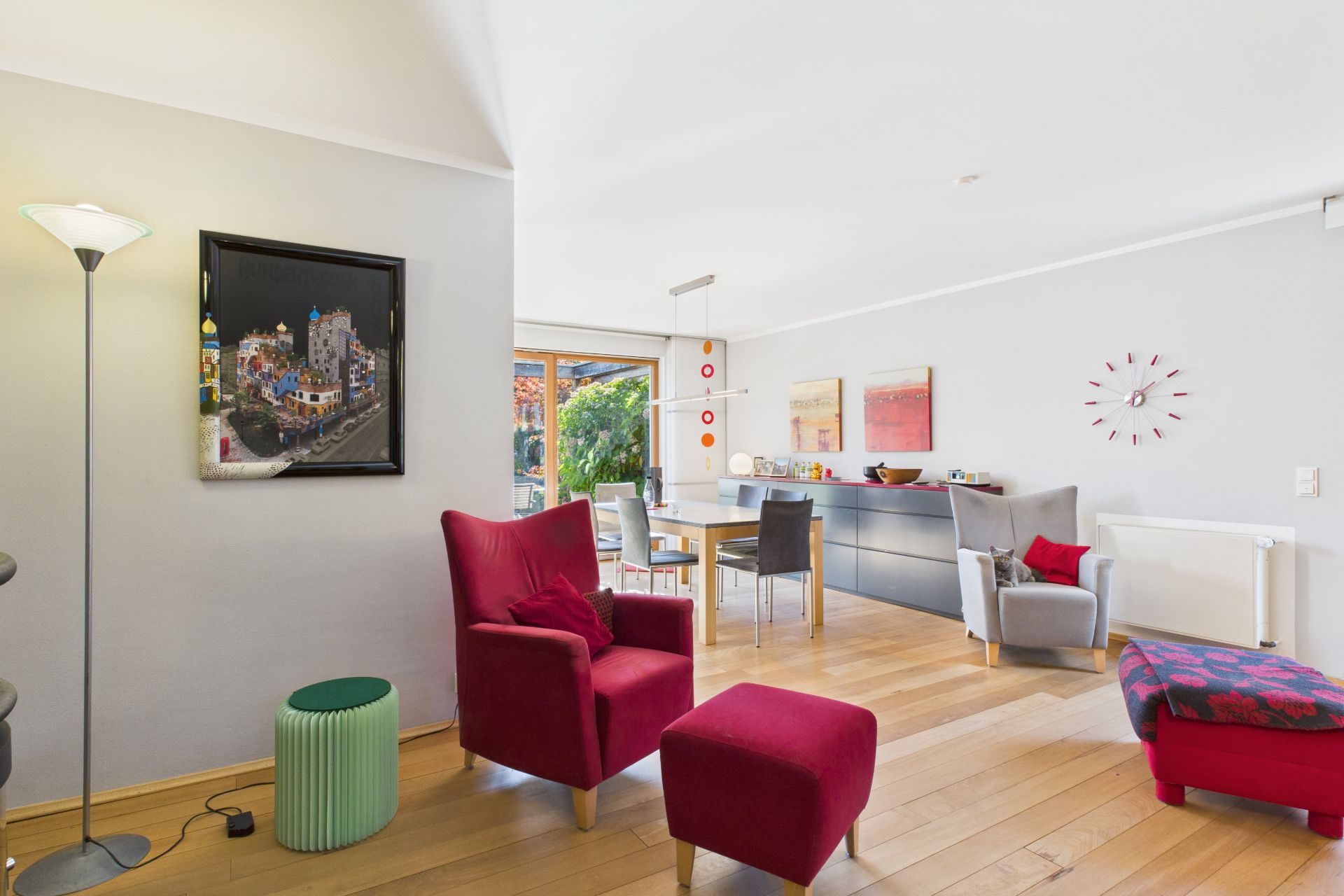 Helles Wohnzimmer mit roten Sesseln, Esstisch, Holzboden und moderner Wanddekoration.