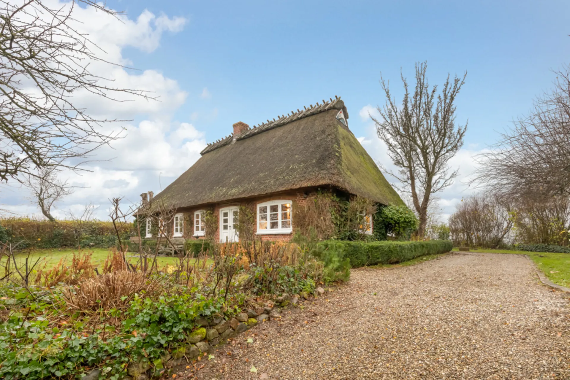 Reetgedecktes Haus mit Garten, Kiesweg und Büschen an einem bewölkten Tag.