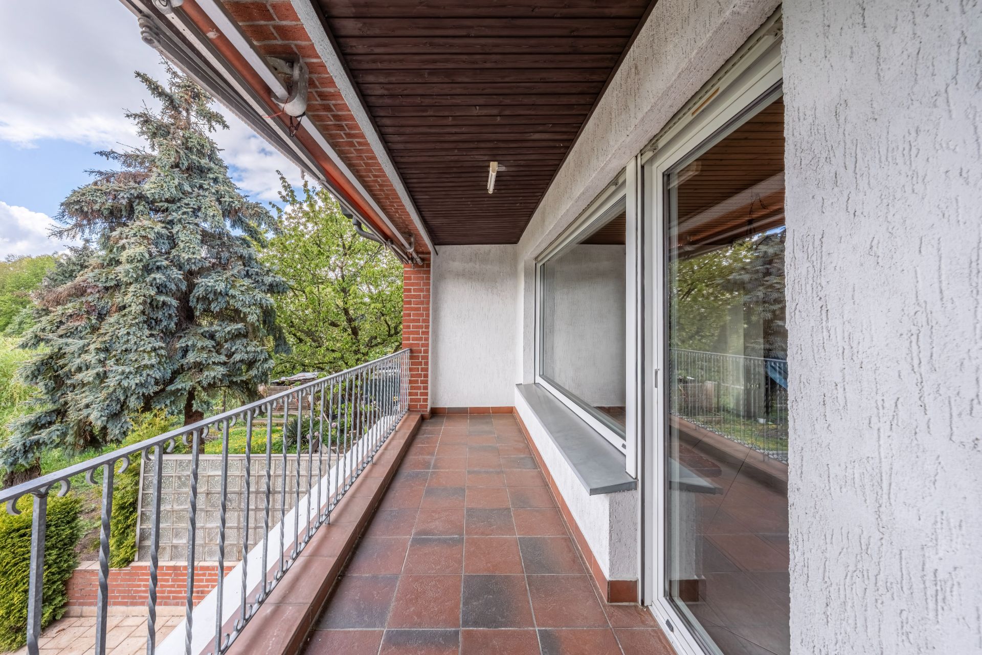 Überdachter Balkon mit Fliesenboden, Metallgeländer und Blick auf Bäume.
