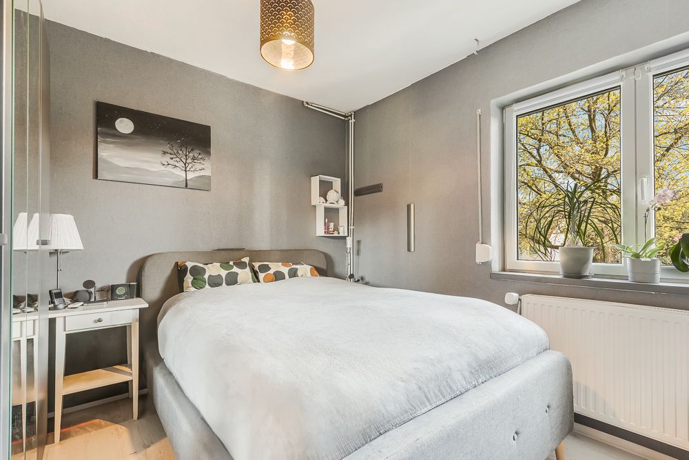 Helles Schlafzimmer mit Doppelbett, Nachttisch, Wandbild und Fenster mit Pflanzen.