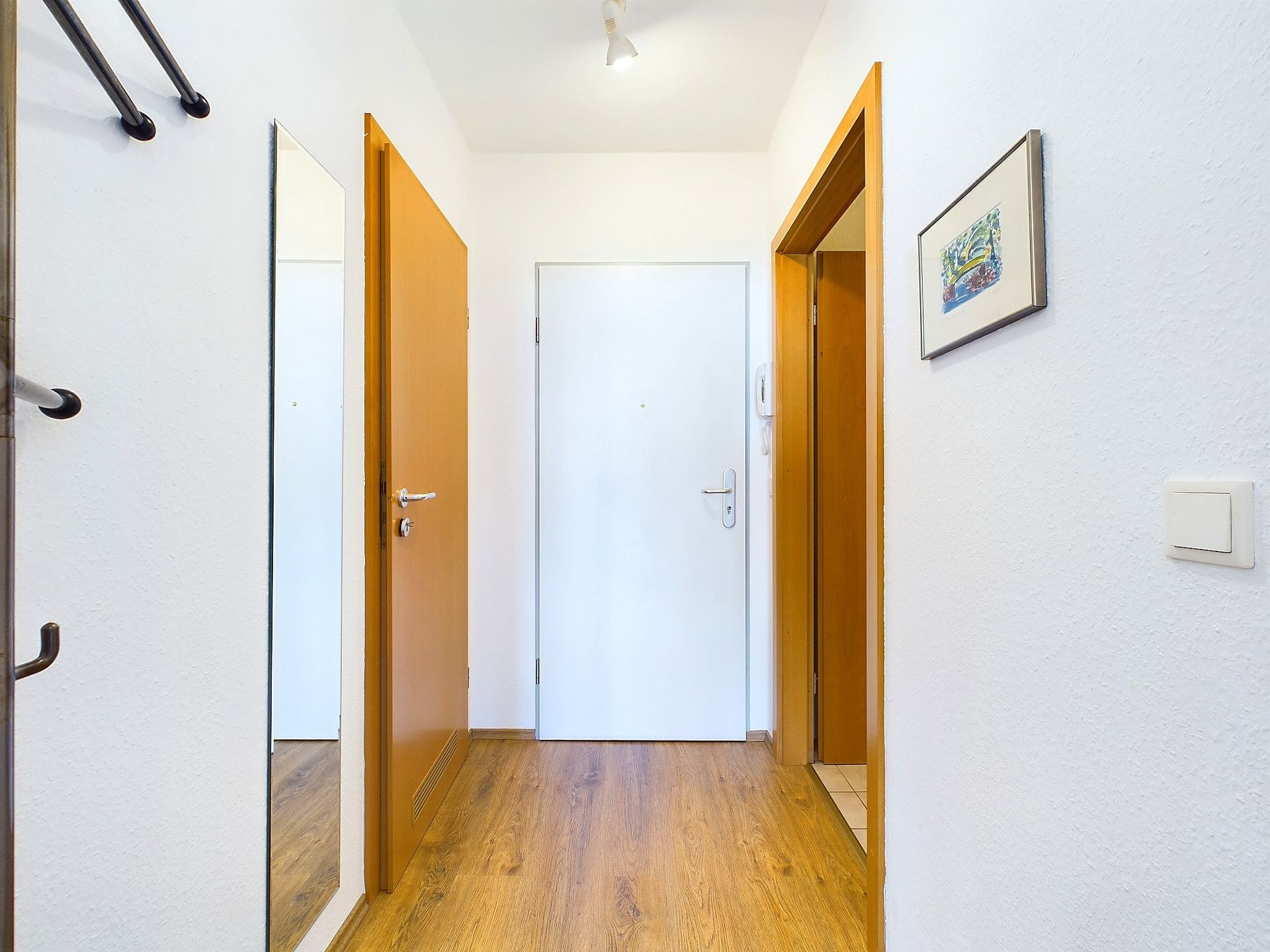 Heller Flur mit Holzboden, weißer Tür, Spiegel und Wandbild.