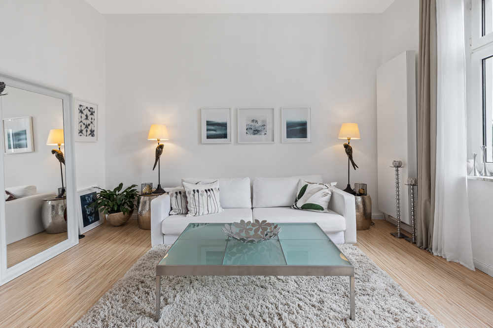 Helles Wohnzimmer mit weißem Sofa, Glastisch, Teppich und Wandlampen neben Bildern an der Wand.