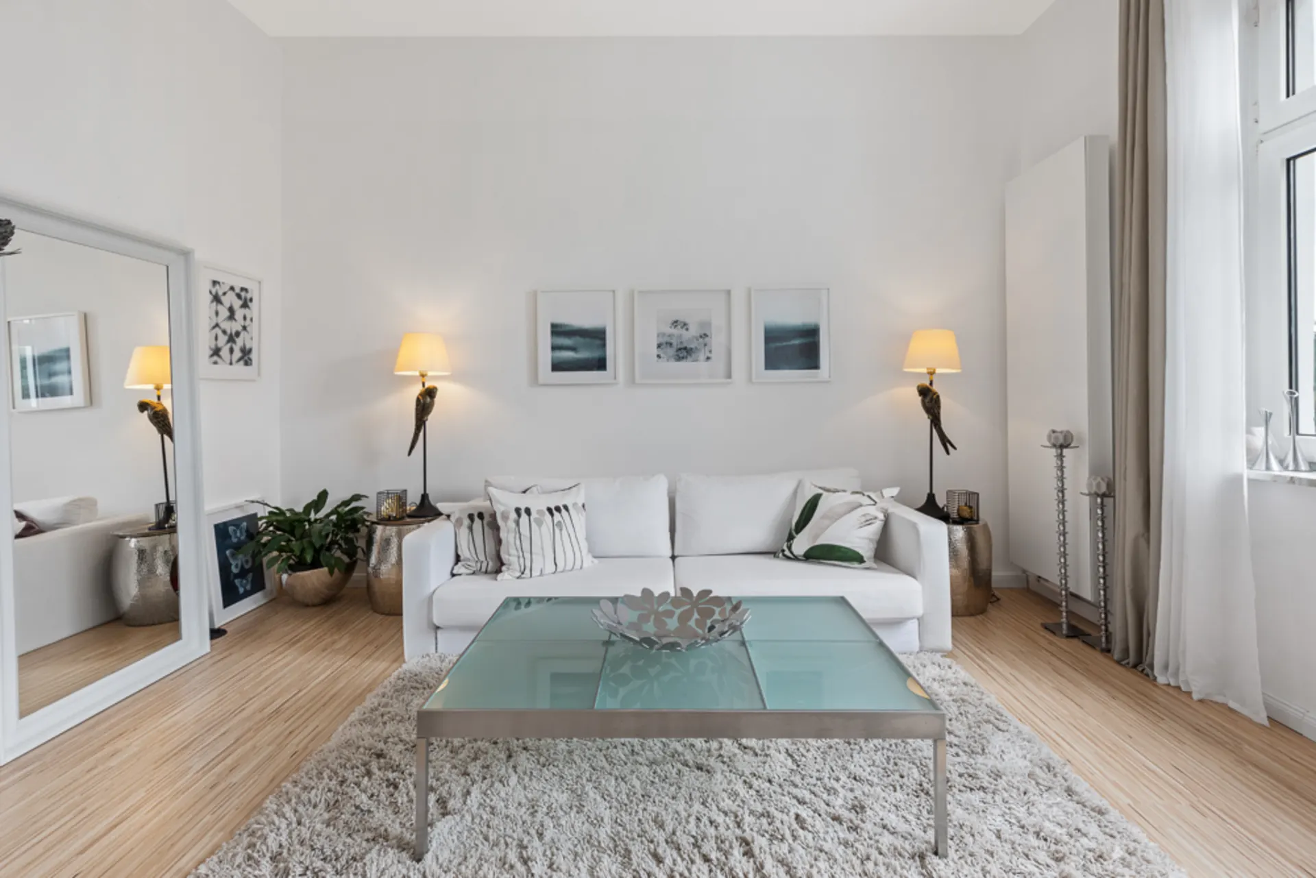 Helles Wohnzimmer mit weißem Sofa, Glastisch, Teppich und Wandlampen neben Bildern an der Wand.