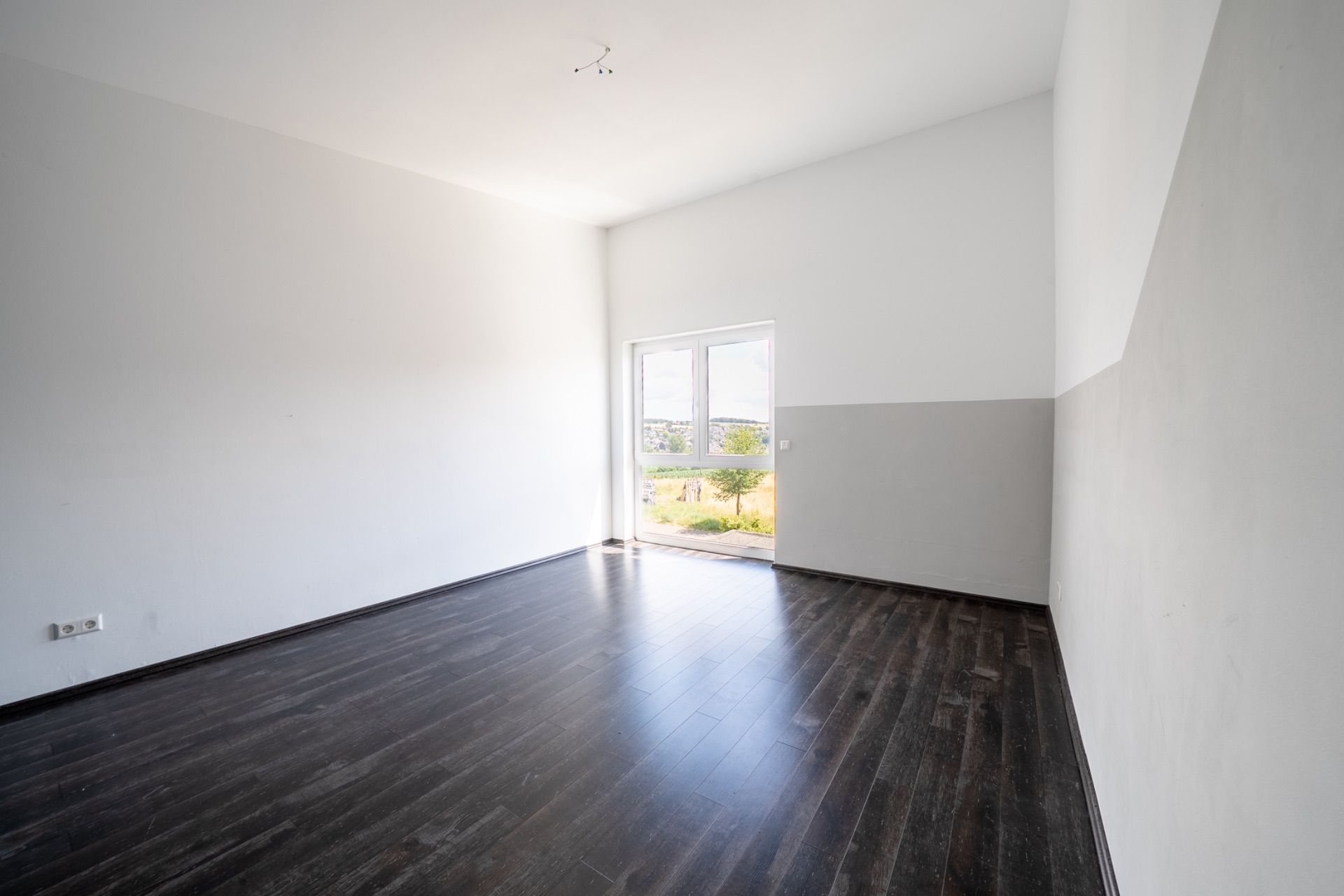 Leerer Raum mit dunklem Holzboden und weiß gestrichenen Wänden, Fenster mit Blick ins Freie.
