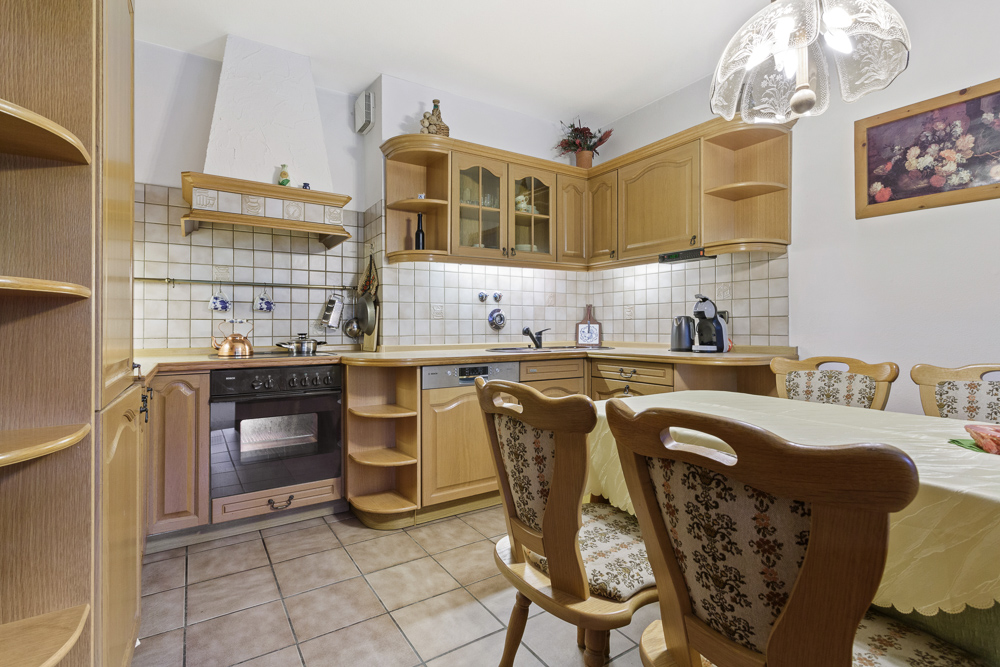 Helle Küche mit Holzmöbeln, Esstisch und vier Stühlen, Fliesenboden und Deckenlampe.
