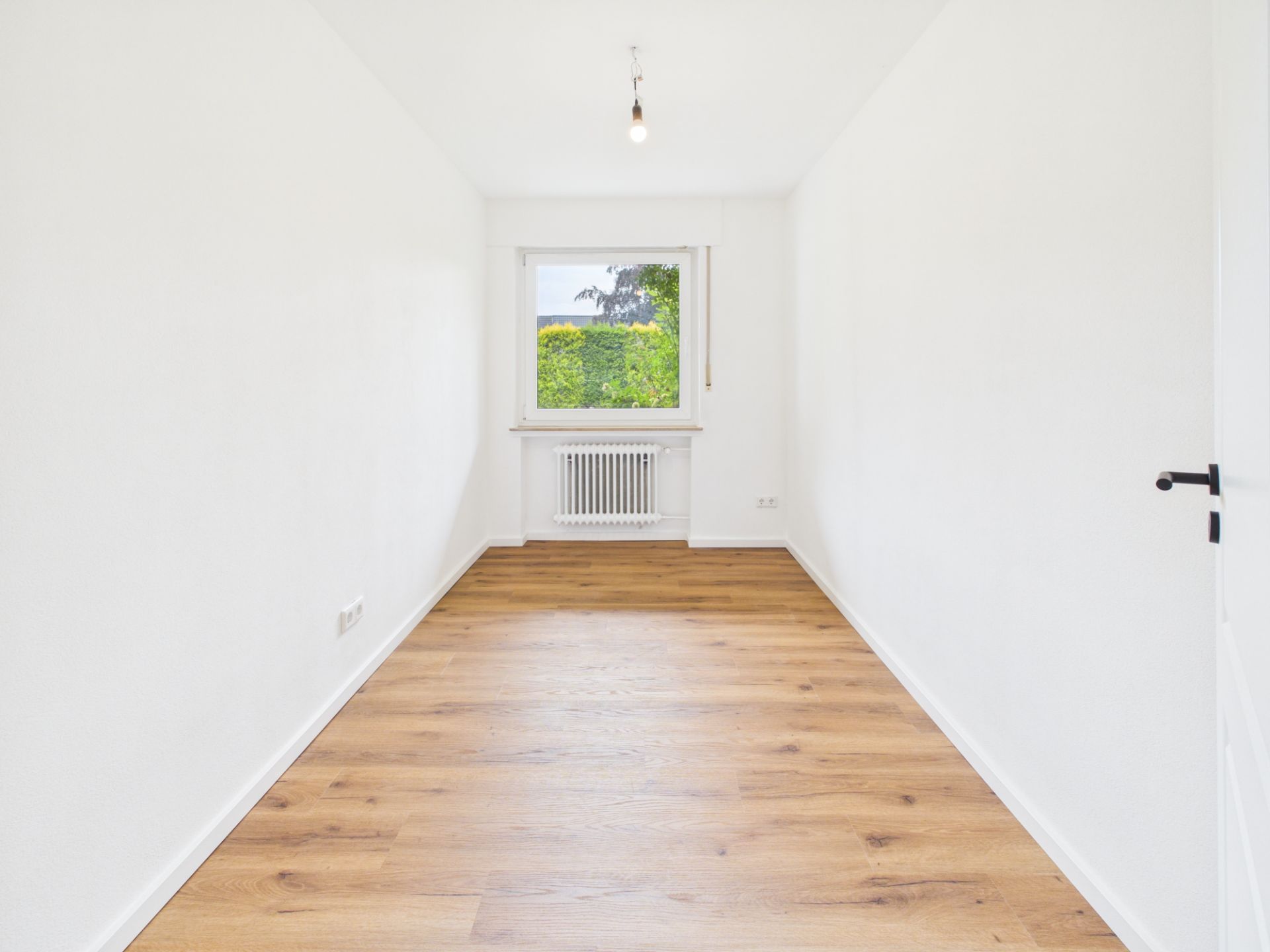 Heller, leerer Raum mit Holzfußboden, weißer Wand und Fenster mit Blick ins Grüne.