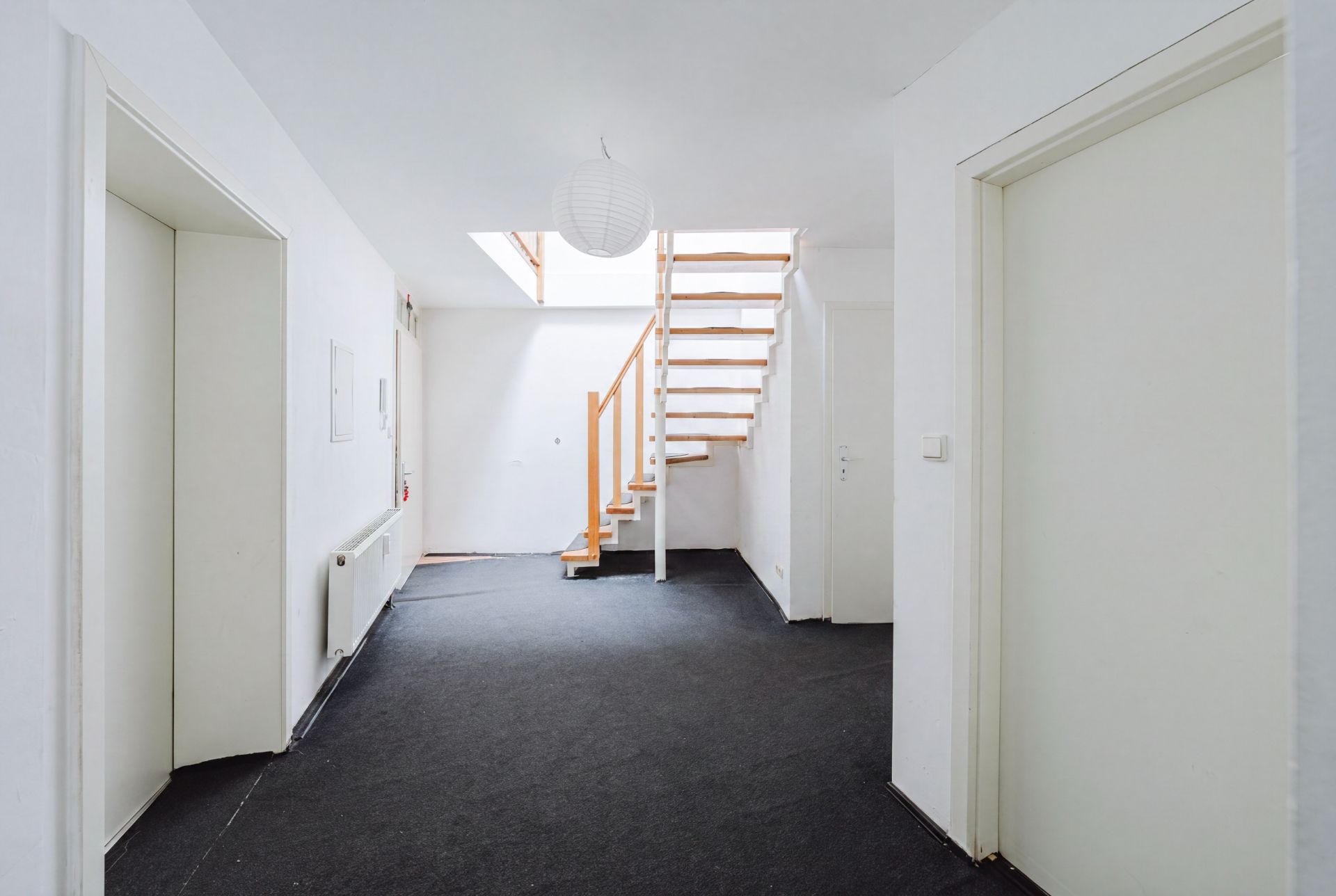 Heller Flur mit schwarzem Teppich, weißen Wänden und einer Holztreppe nach oben.