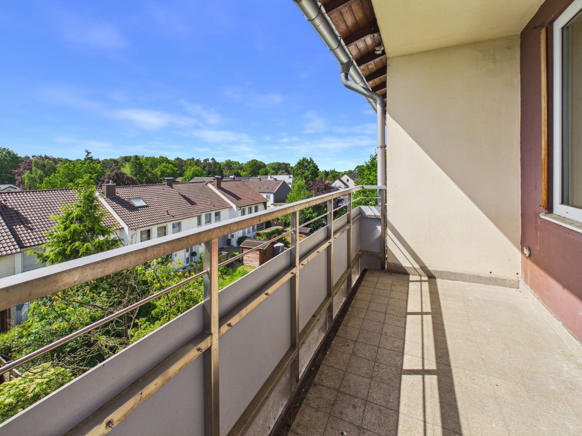 Balkon mit Metallgeländer, Blick auf Nachbarhäuser und grüne Bäume bei klarem Himmel.