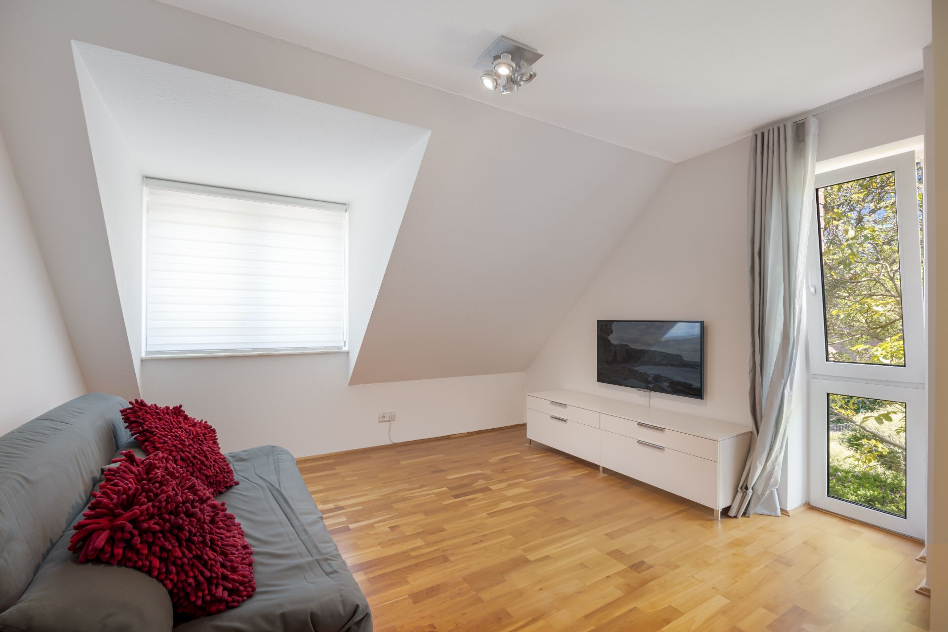 Wohnzimmer mit Sofa, roten Kissen, TV auf weißem Schrank und großen Fenstern mit Vorhängen.