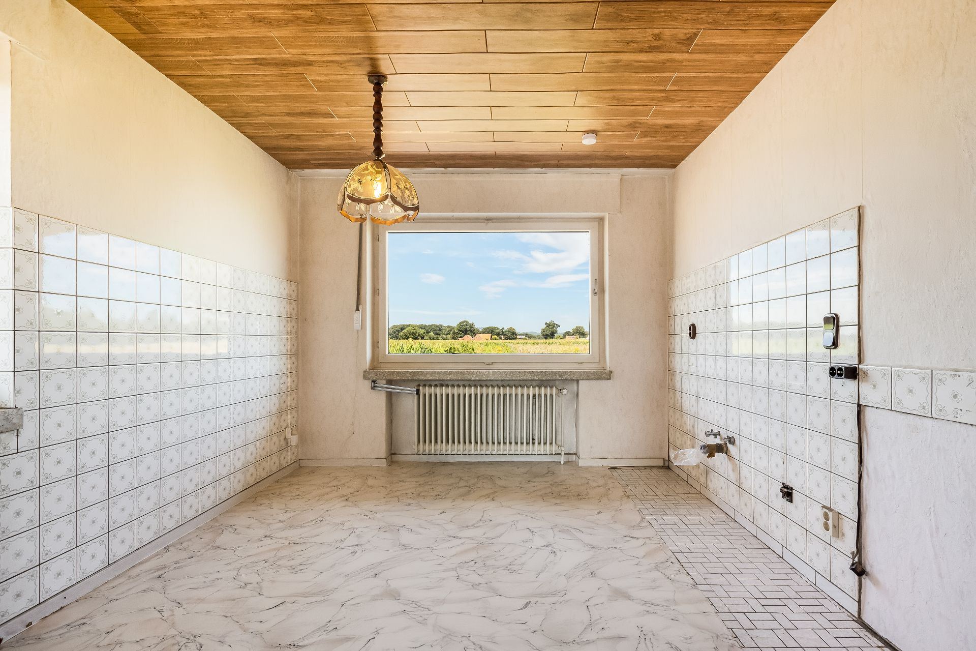 Leerer Raum mit Fliesenwänden, großem Fenster, Heizkörper und Hängelampe an der Decke.