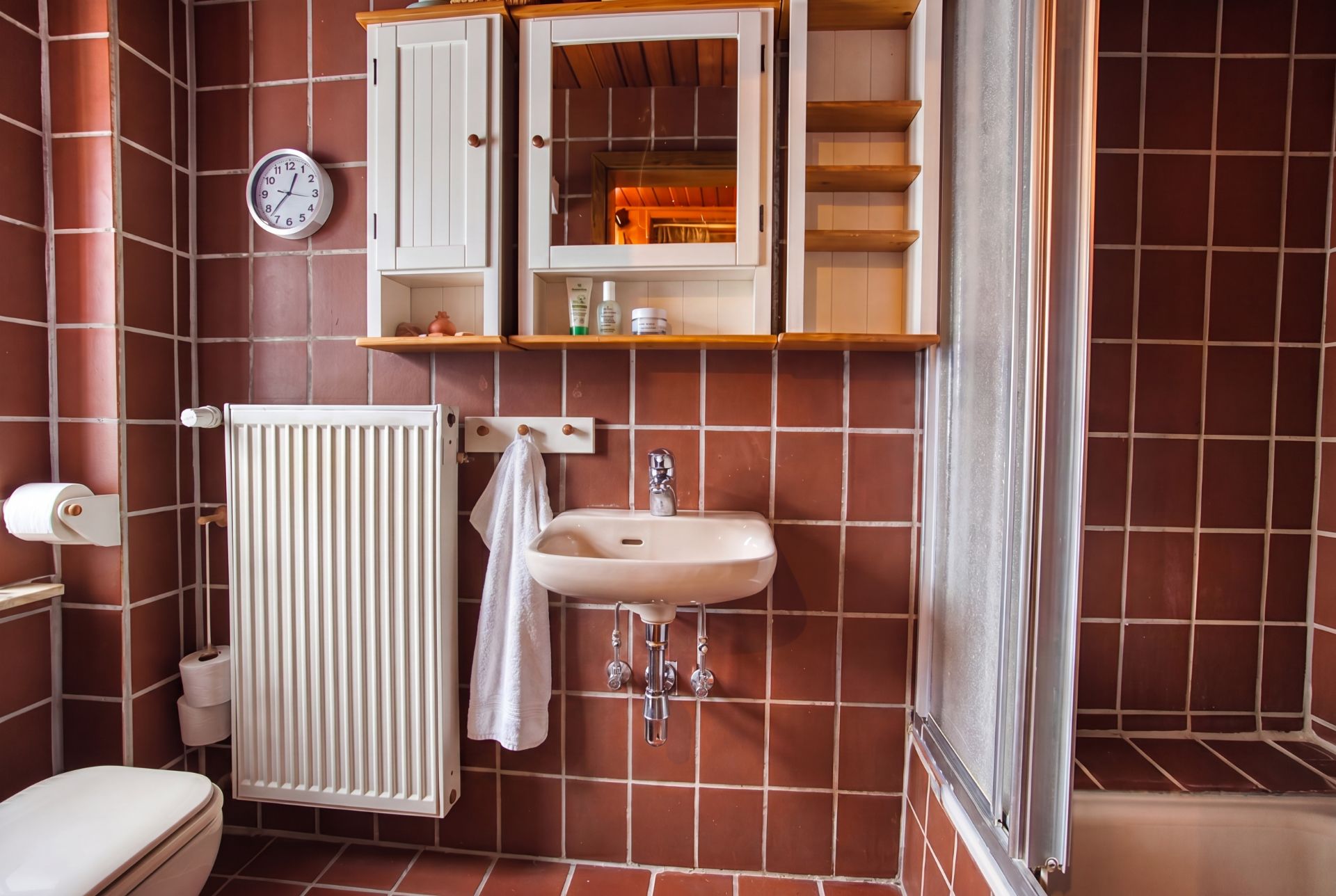 Badezimmer mit braunen Fliesen, Waschbecken, Spiegel, Heizung, WC und Badewanne.
